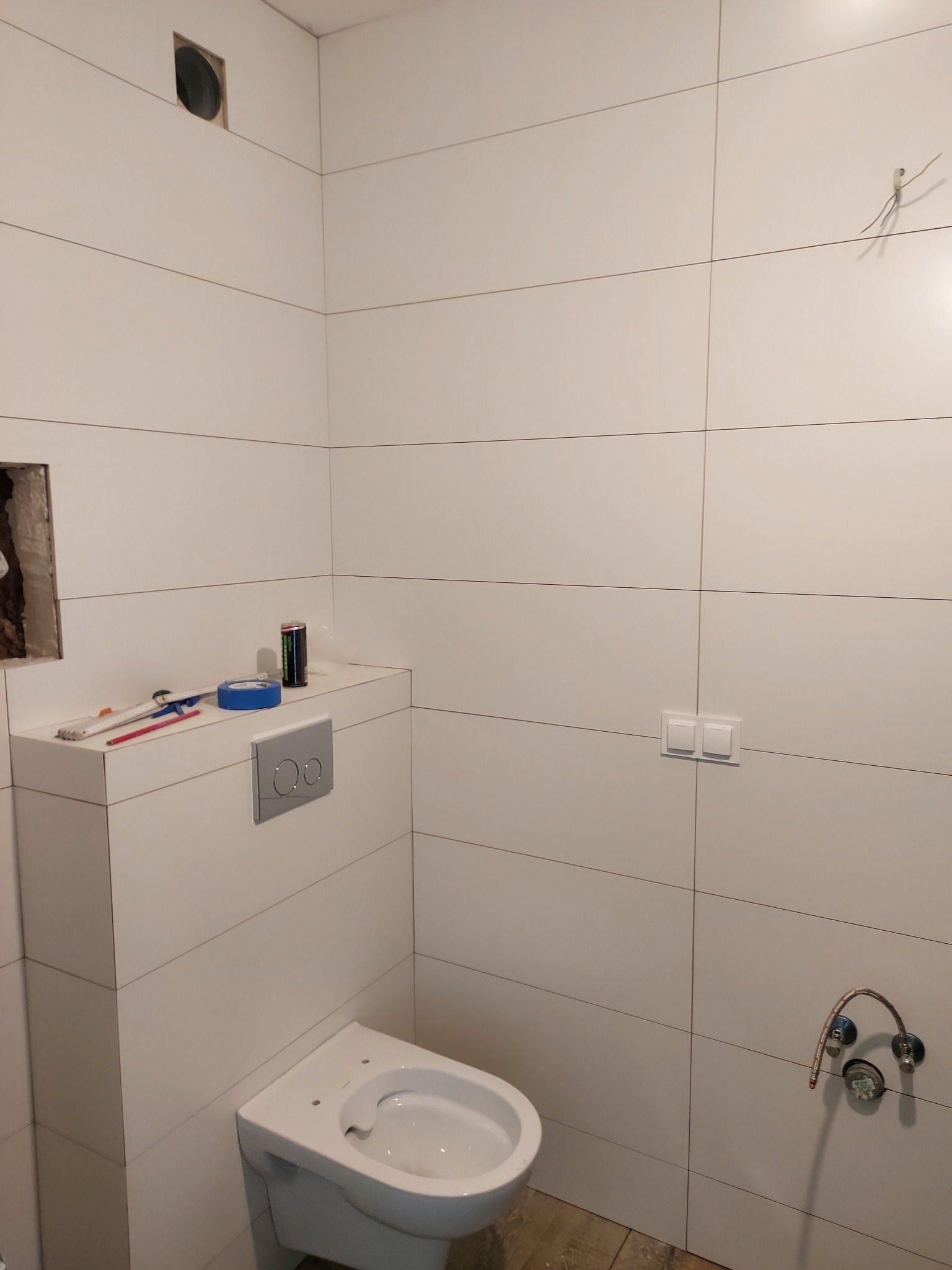 Biała łazienka w trakcie wykańczania, widoczny częściowo zamontowany sedes podtynkowy, białe płytki ścienne, wystające kable, otwór wentylacyjny oraz narzędzia i materiały budowlane na zabudowie...