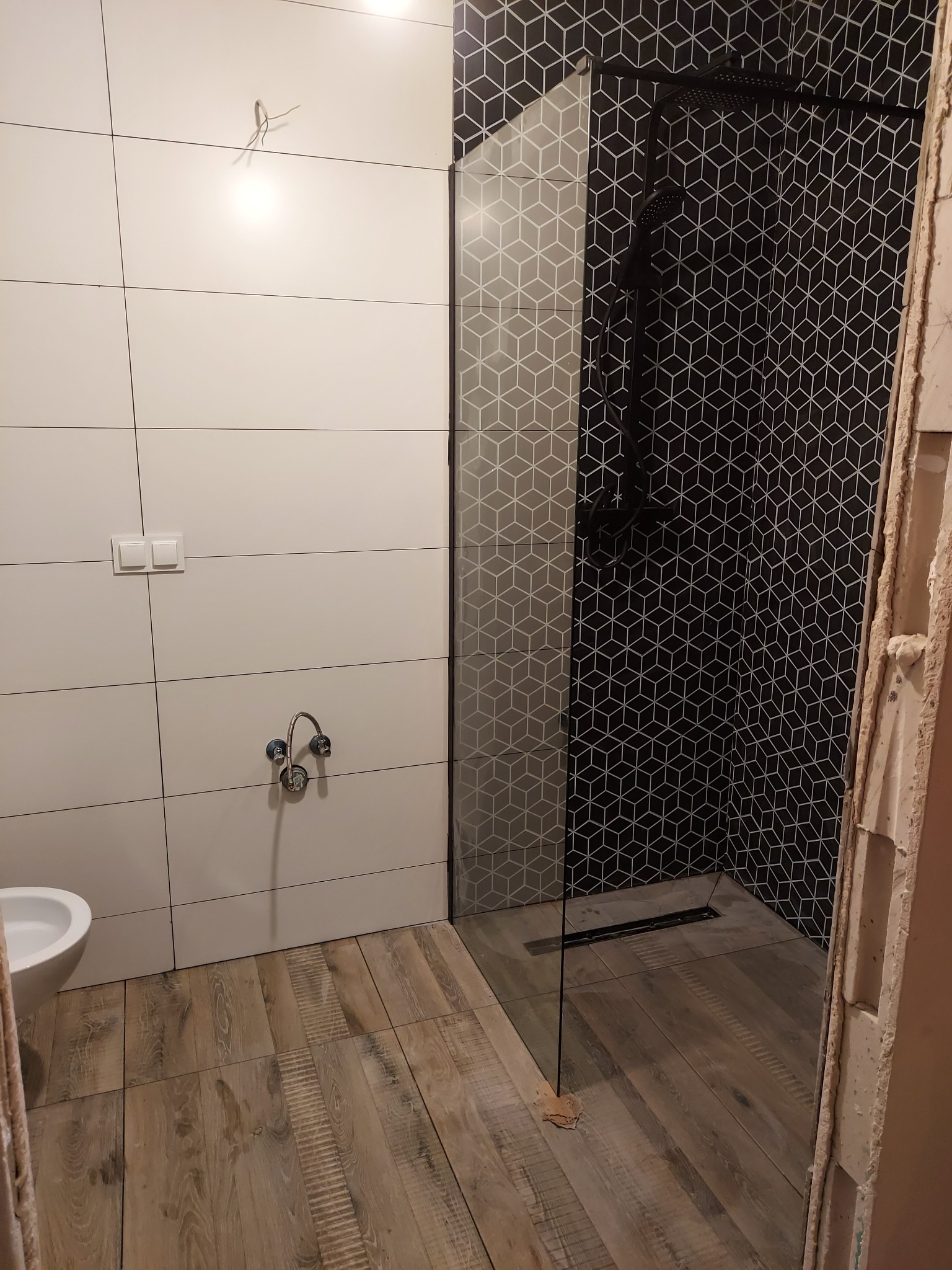 Łazienka w trakcie remontu: białe płytki ścienne, drewnopodobna podłoga, kabina prysznicowa ze wzorem geometrycznym, widoczne elementy instalacji wodnej i niedokończone wykończenia.