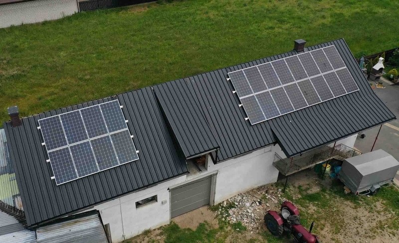Widok z góry na budynek gospodarczy z dwiema instalacjami paneli słonecznych na dachu z blachy trapezowej, obok stoi czerwony traktor.