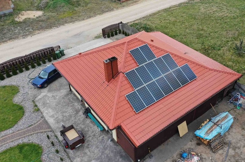 Widok z góry na dom z czerwoną dachówką i panelami słonecznymi na dachu, obok komina. Na podwórku zaparkowany niebieski samochód, przyczepka i materiały budowlane pod plandeką.