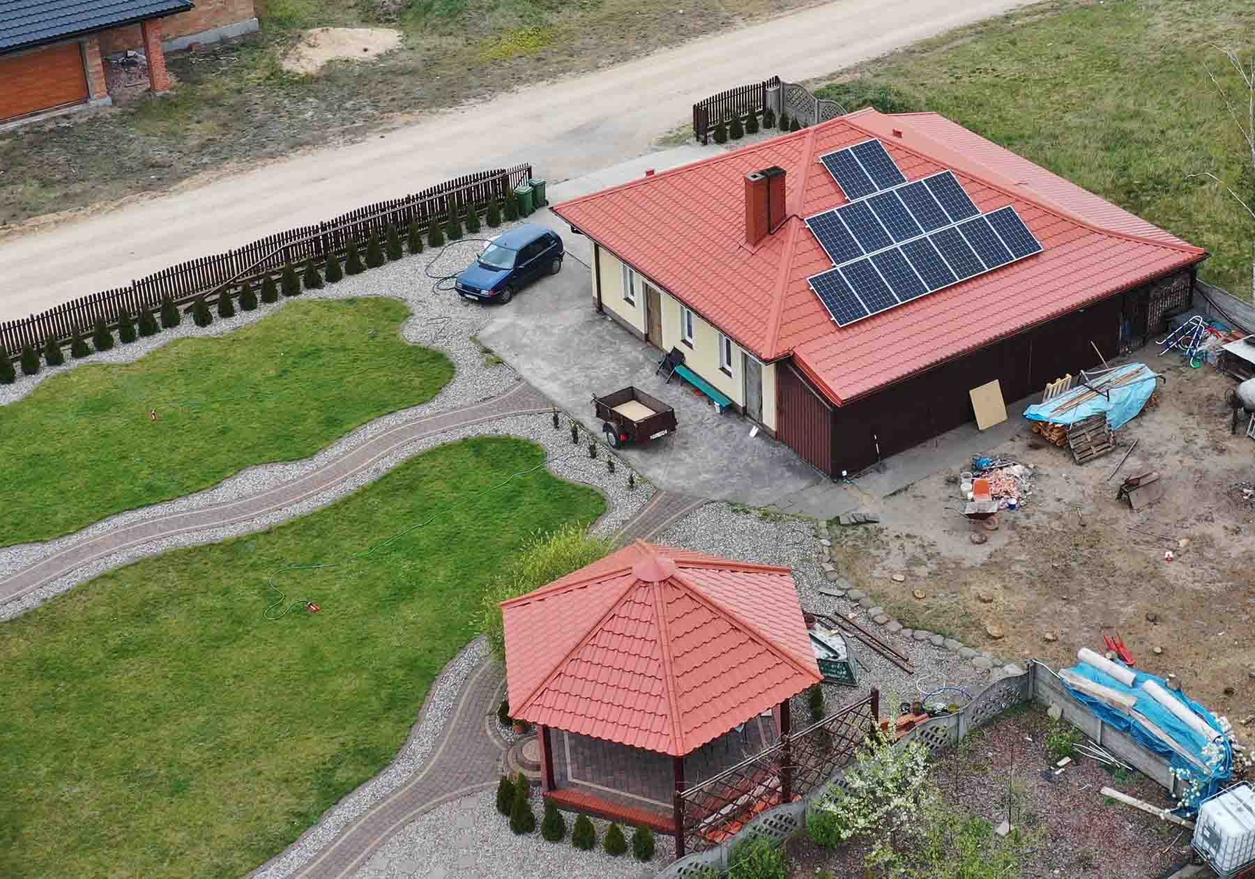 Widok z góry na dom jednorodzinny z czerwonym dachem i zainstalowanymi panelami słonecznymi. Obok domu zaparkowany samochód i stoi przyczepka. W tle widoczna zadbana działka z altaną.