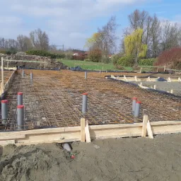 Zbrojenie fundamentów pod dom jednorodzinny z widocznymi pionowymi rurami kanalizacyjnymi i drewnianym szalunkiem, w tle zielony krajobraz z drzewami i błękitne niebo.