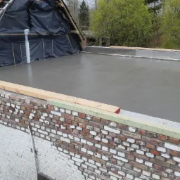 Wylewka betonowa na dachu budynku z ceglaną ścianą szczytową i zabezpieczeniem folią przed dalszymi pracami dekarskimi.