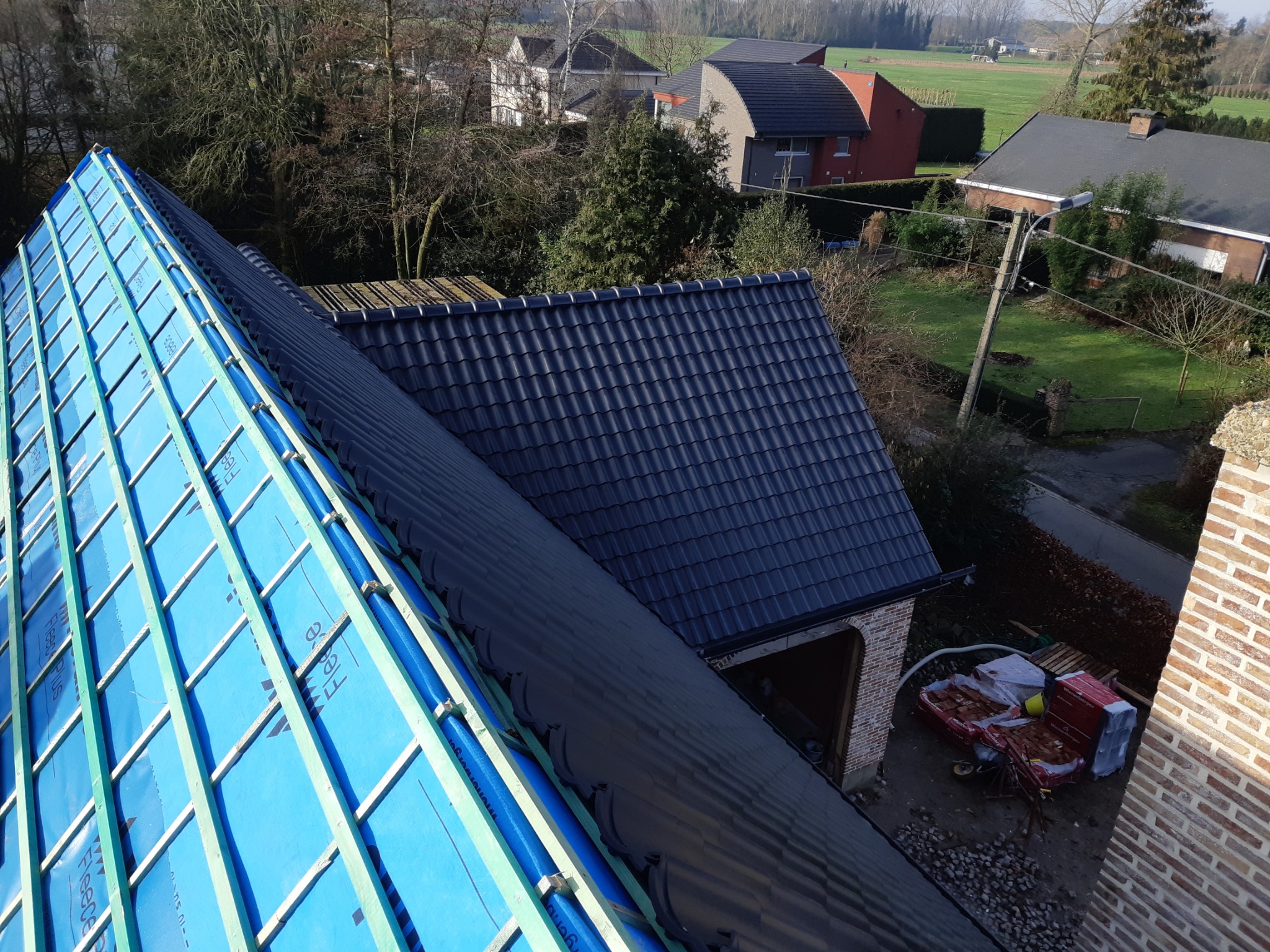 Widok z góry na fragment dachu z nową więźbą pokrytą niebieską membraną izolacyjną, obok fragment dachu pokrytego czarną dachówką, w tle zabudowania mieszkalne i pola.