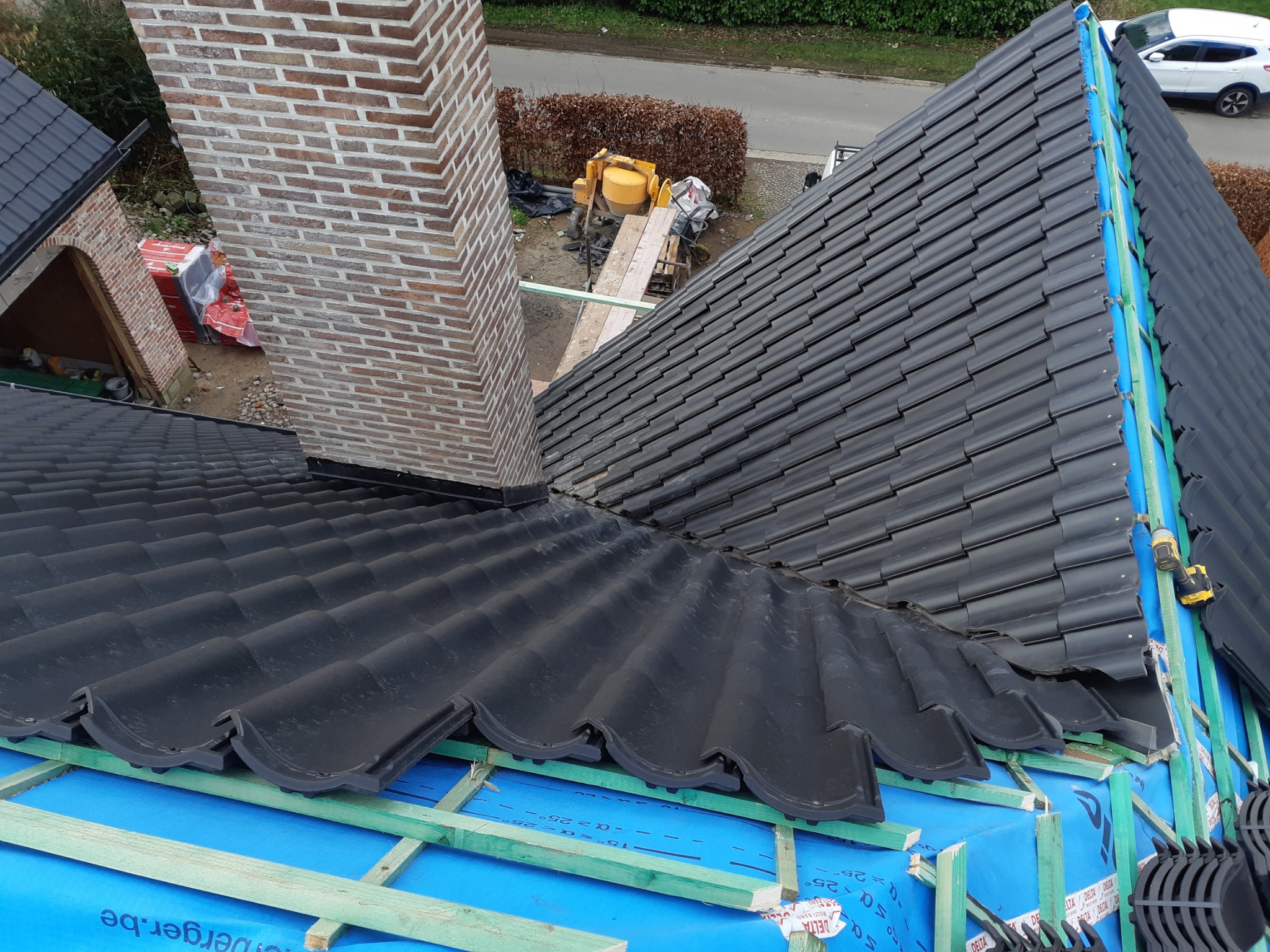Czarny dach z dachówki ceramicznej w trakcie układania, widok z góry, komin z cegły, na podkładzie niebieska membrana i drewniana konstrukcja nośna, w tle betoniarka i zaparkowany samochód.