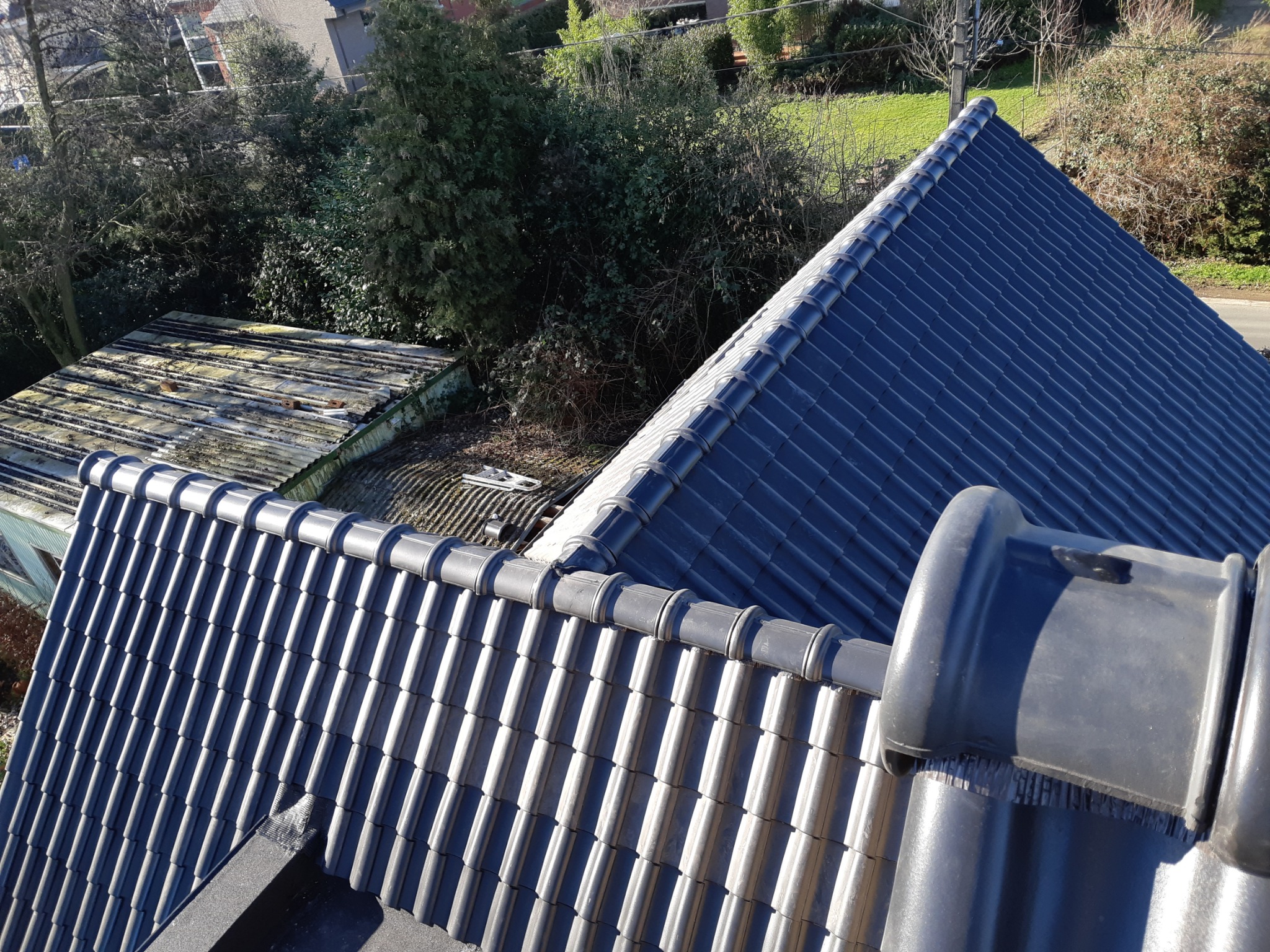 Nowy, ciemny dach z ceramicznej dachówki karpiówki, widok z góry na tle zieleni i starego, zardzewiałego dachu.