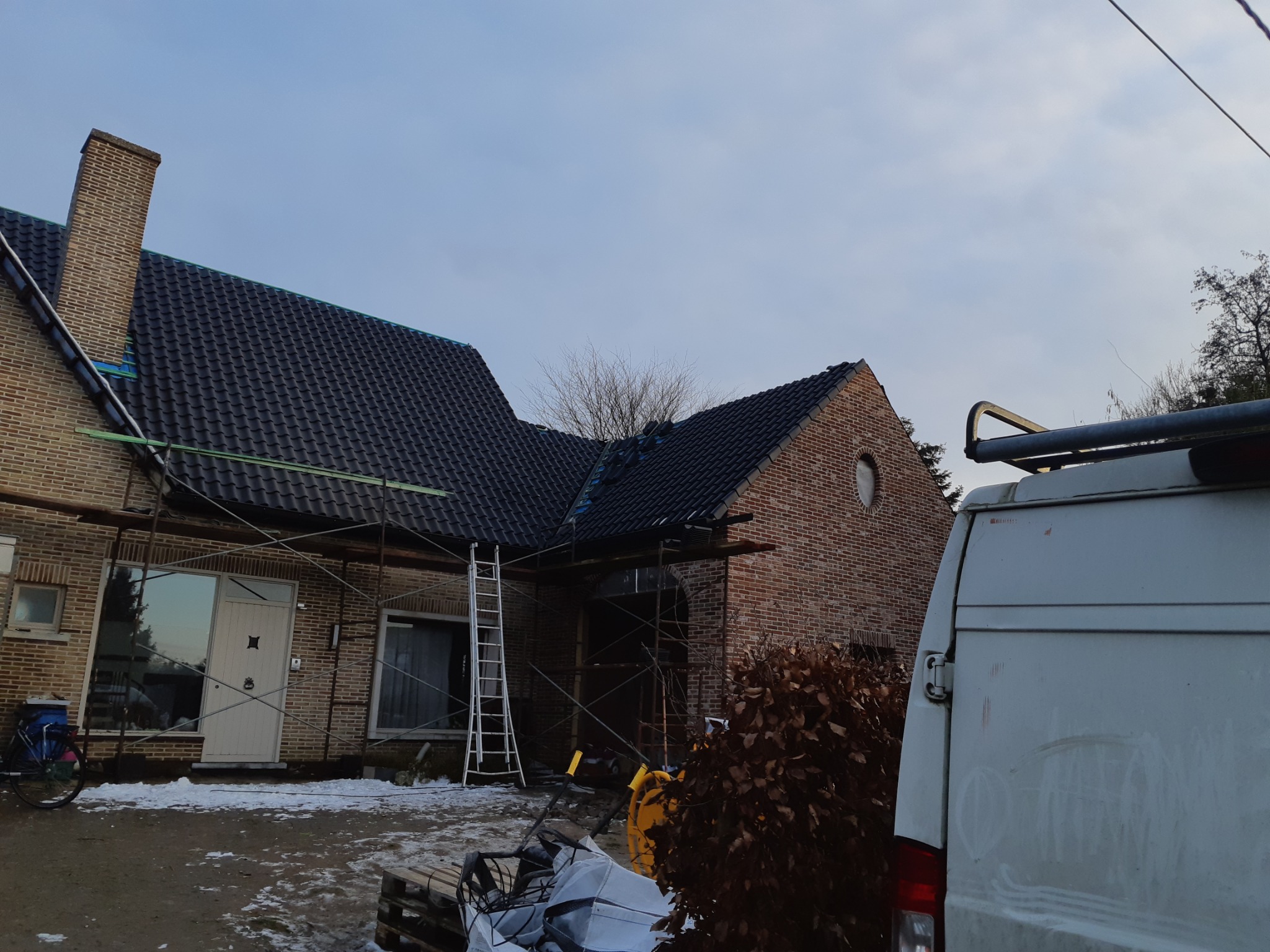 Dom z cegły, częściowo pokryty nowym, czarnym dachem, otoczony rusztowaniami podczas prac, widoczny samochód dostawczy i narzędzia.