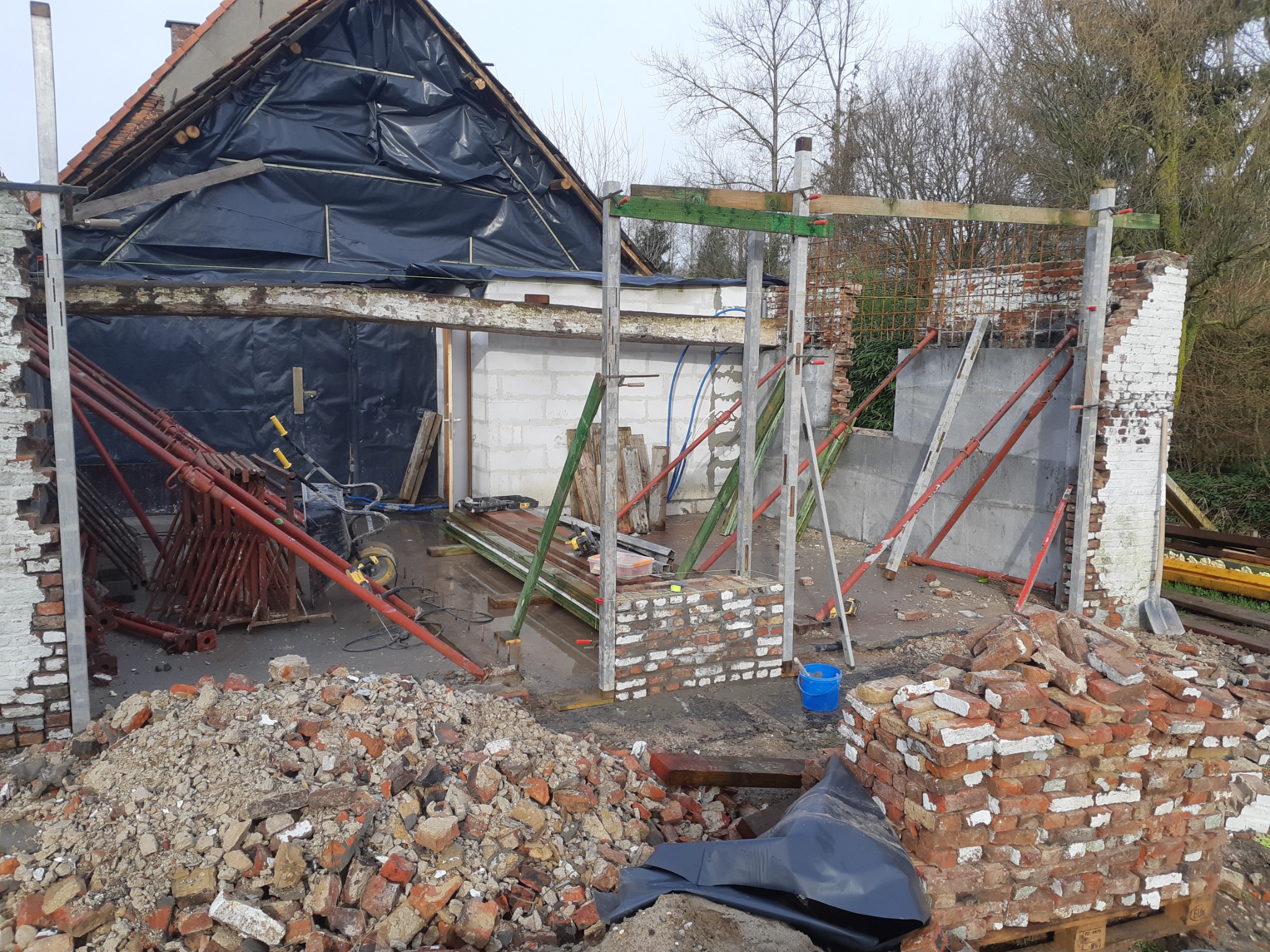 Rozbiórka i przygotowanie do budowy: fragment ściany z cegły, sterta gruzu, elementy konstrukcyjne dachu zabezpieczone folią, podpory budowlane i widoczne narzędzia.