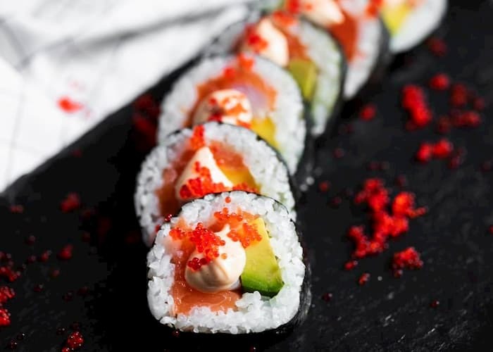 Rząd rolek sushi z łososiem, awokado i sosem majonezowym, posypanych czerwonym kawiorem, ułożonych na czarnym talerzu z białym ręcznikiem w tle.