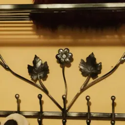 Kuty metalowy wieszak na ubrania z półką, zdobiony motywem liści i kwiatów, zawieszony na żółtej ścianie.