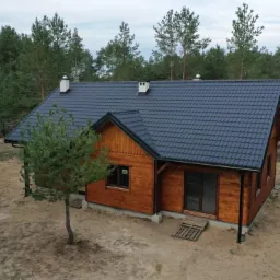 Gotowy, parterowy dom z drewna, z ciemną dachówką i dwoma kominami, otoczony lasem sosnowym, widok z góry.