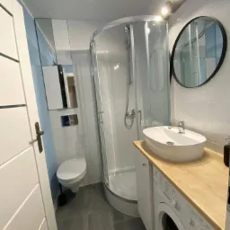 Nowoczesna łazienka z kabiną prysznicową, umywalką nablatową na drewnianym blacie, pralką w zabudowie, szarą podłogą i okrągłym lustrem, oświetlona listwą LED.