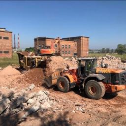 Wyburzenia Śląsk - Pomarańczowa ładowarka kołowa Husqvarna transportuje gruz, w tle koparka i budynki z czerwonej cegły, na pierwszym planie stos gruzu.
