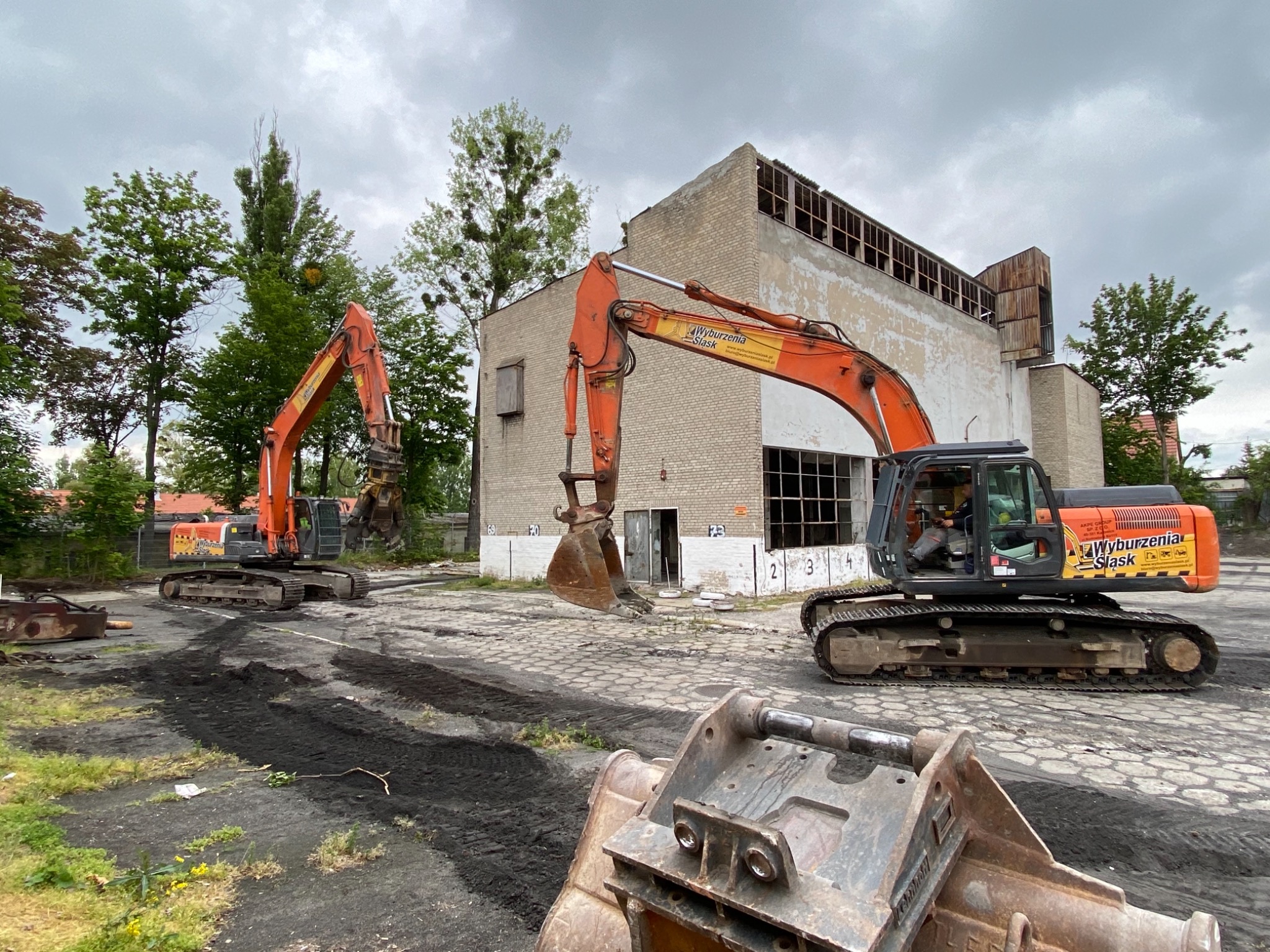 Dwie pomarańczowe koparki gąsienicowe na terenie rozbiórki ceglanego budynku z pustymi otworami okiennymi, widoczne ślady gąsienic na ziemi.