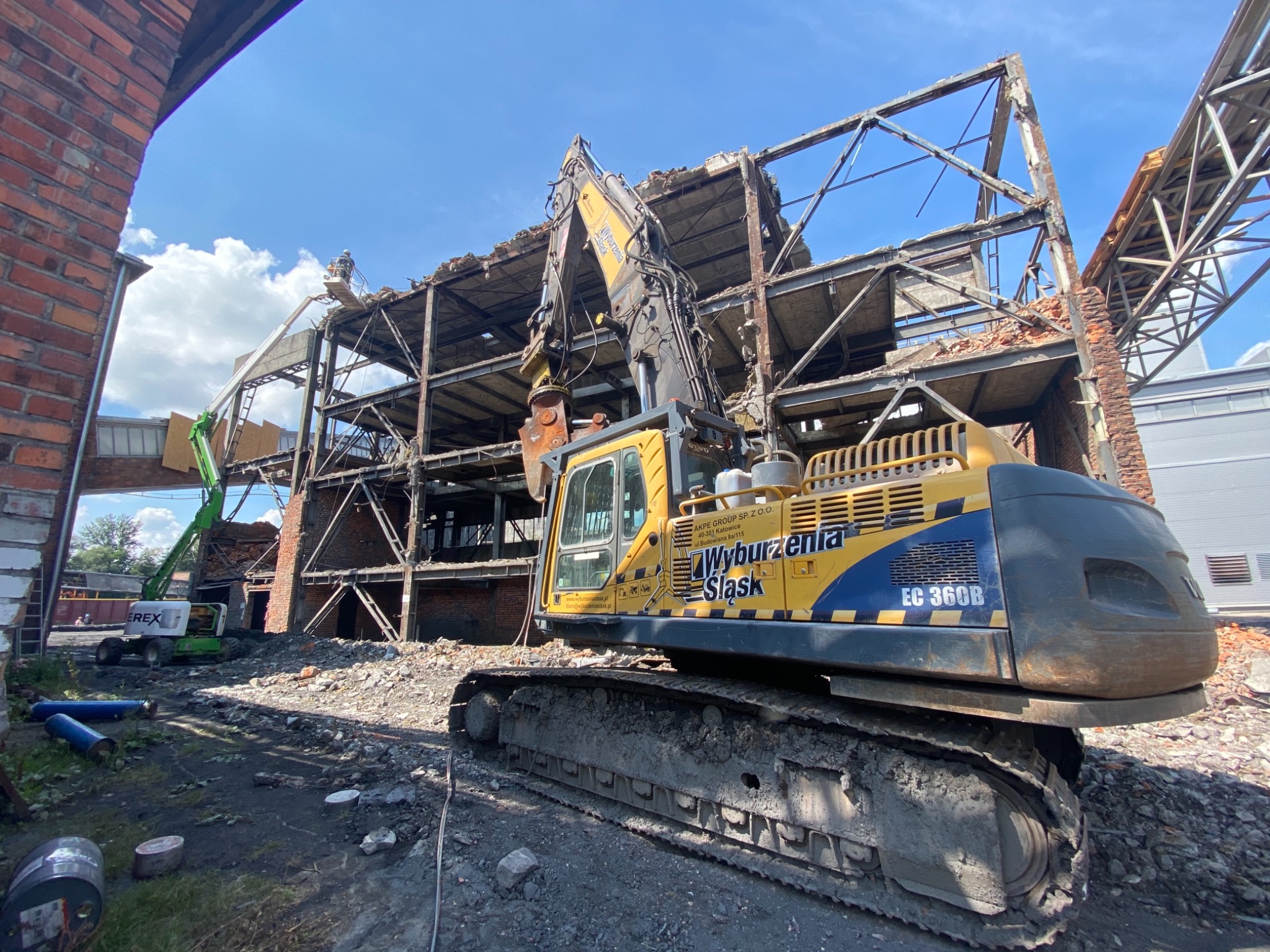 Żółto-niebieska koparka gąsienicowa Wyburzenia Śląsk EC 360B na placu rozbiórki, w tle częściowo zburzony, wielopiętrowy budynek z cegły i stali, oraz zielony podnośnik koszowy.