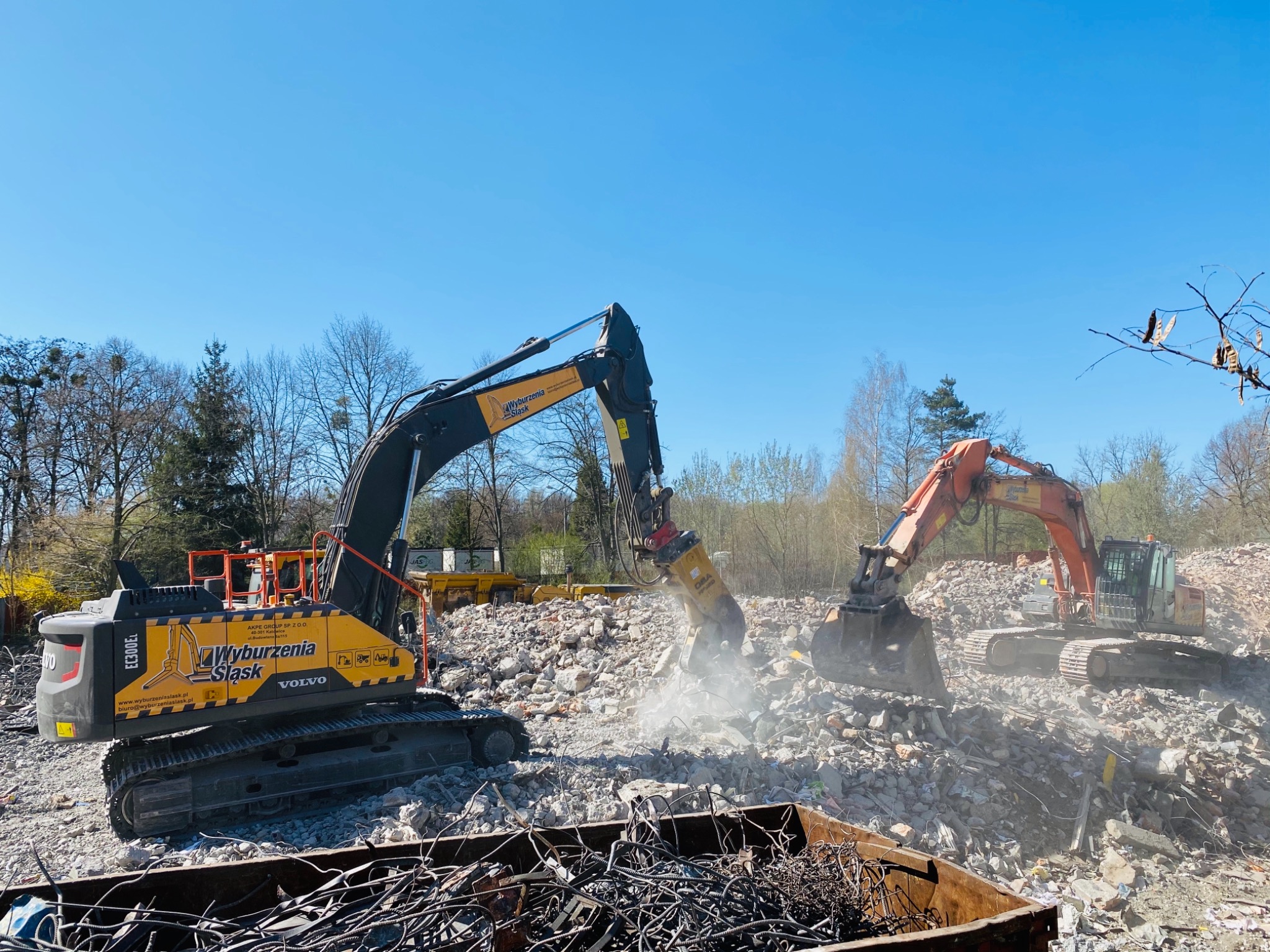 Dwa ciężkie, żółto-czarne i pomarańczowe koparki na gruzowisku, jedna z młotem hydraulicznym kruszy beton, w tle drzewa bez liści i błękitne niebo.