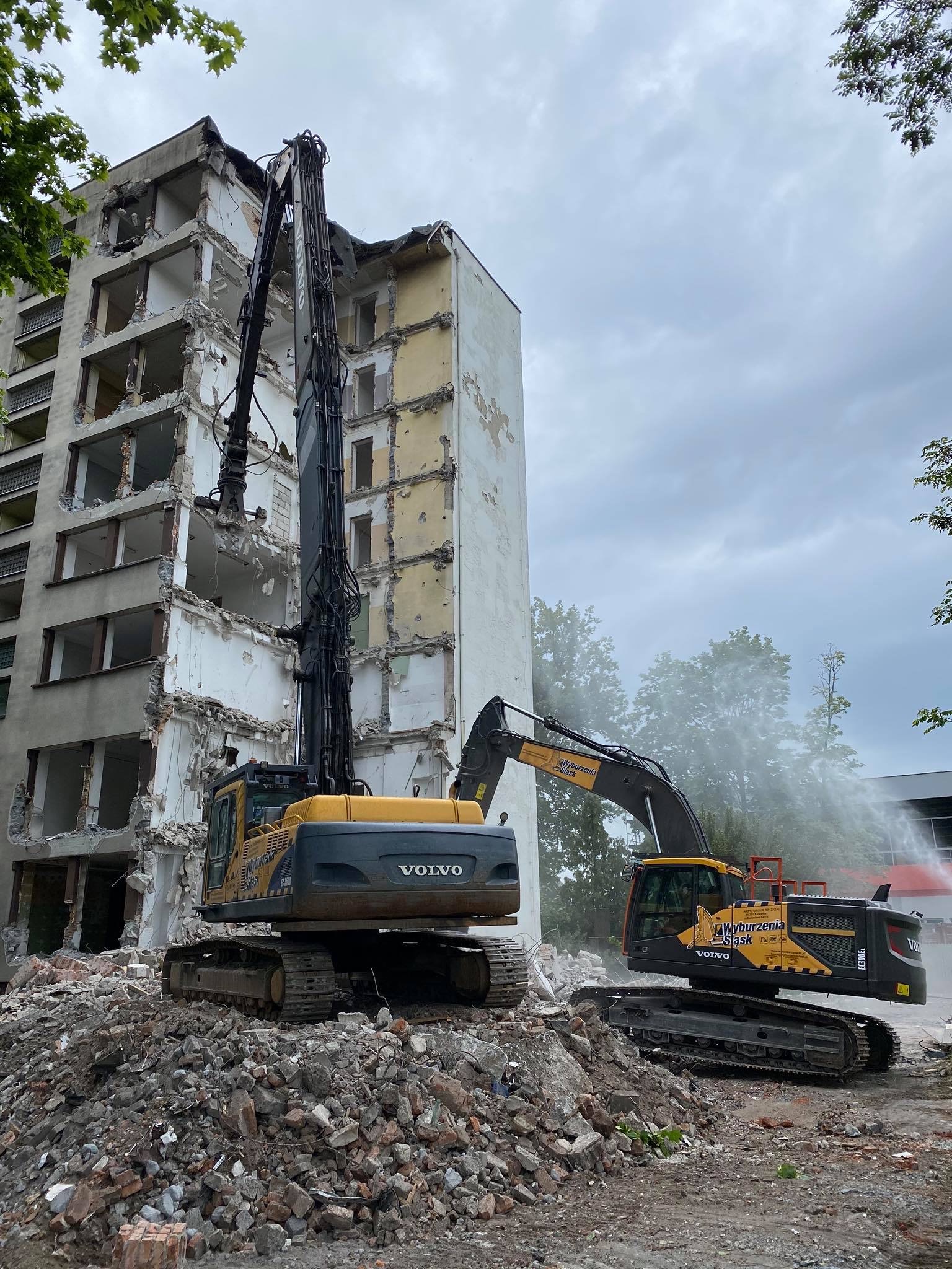 Dwa żółte i czarne koparki Volvo na gruzowisku podczas rozbiórki budynku mieszkalnego, jedna z nich spryskuje wodą, aby ograniczyć pylenie.