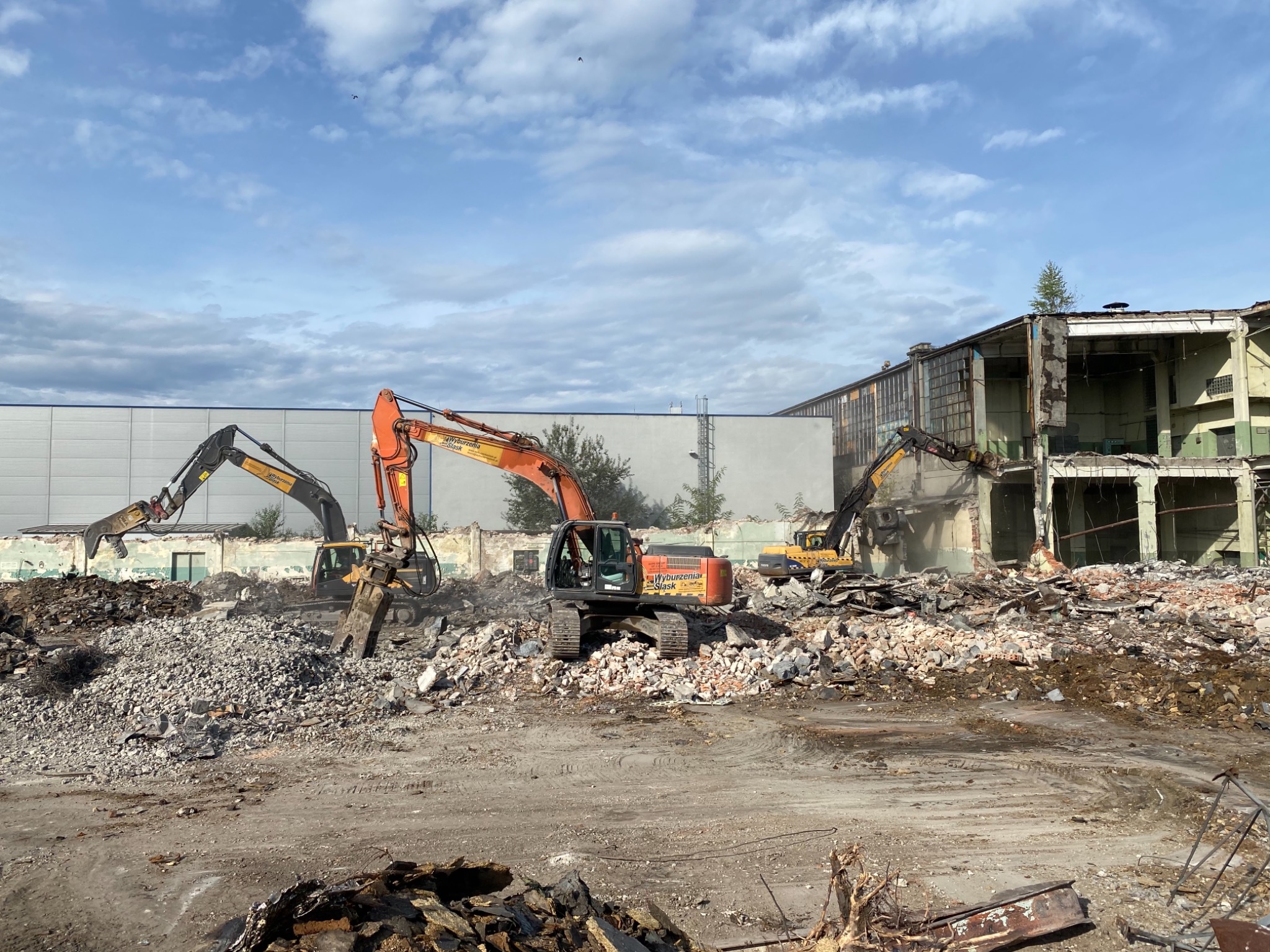 Trzy koparki pracują na placu budowy, rozbierając budynek fabryczny; widoczne gruzy, fragmenty ścian i konstrukcji, a w tle hala przemysłowa pod błękitnym niebem.