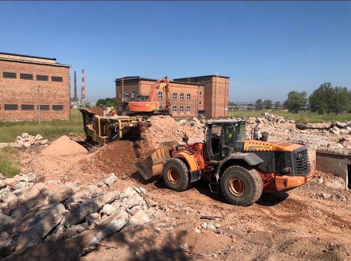 Pomarańczowa ładowarka kołowa Husqvarna transportuje gruz, w tle koparka i budynki z czerwonej cegły, na pierwszym planie stos gruzu.