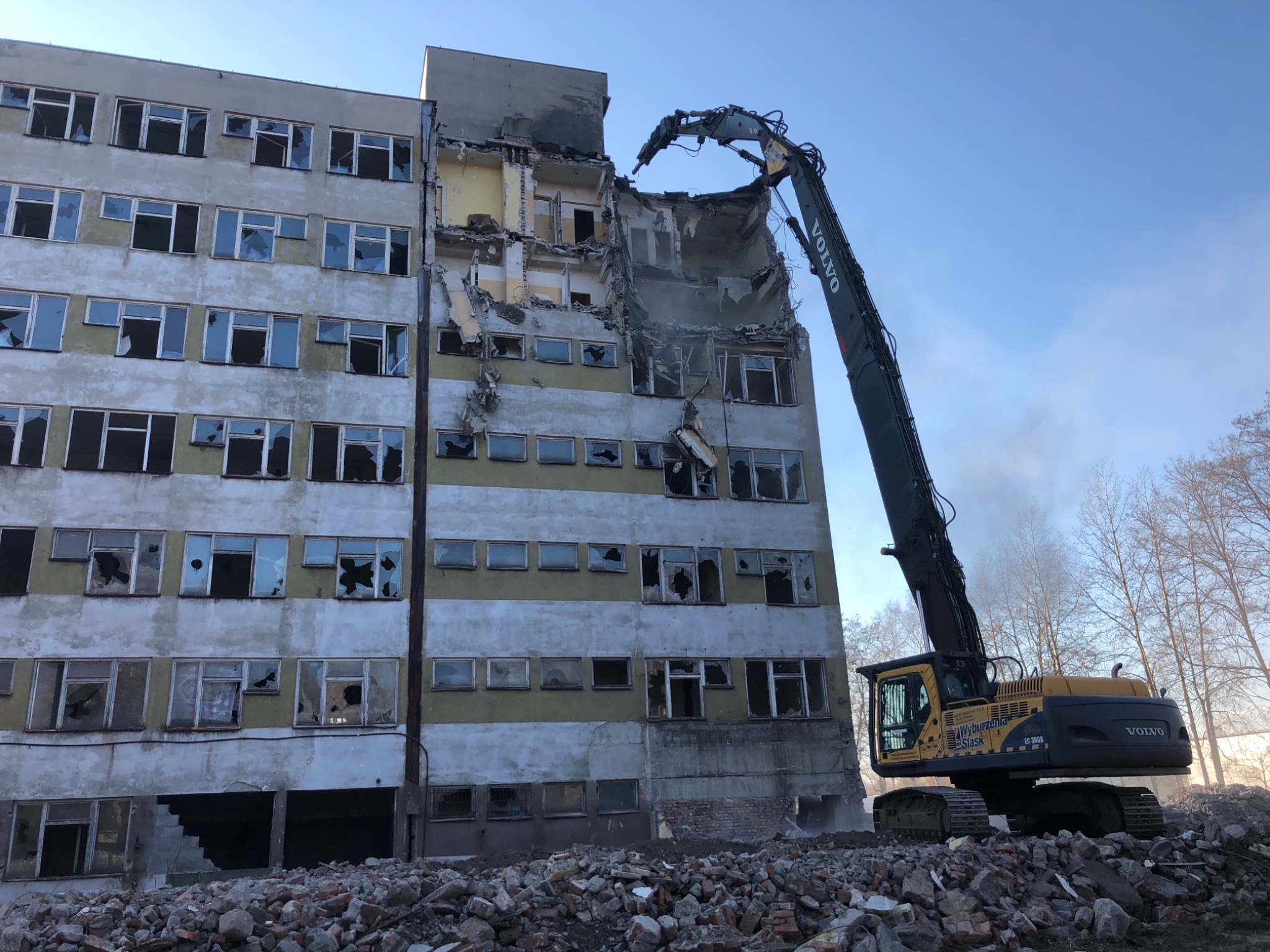 Rozbiórka budynku w Katowicach z użyciem koparki Volvo, widoczne gruzowisko i uszkodzona elewacja budynku z powybijanymi oknami.