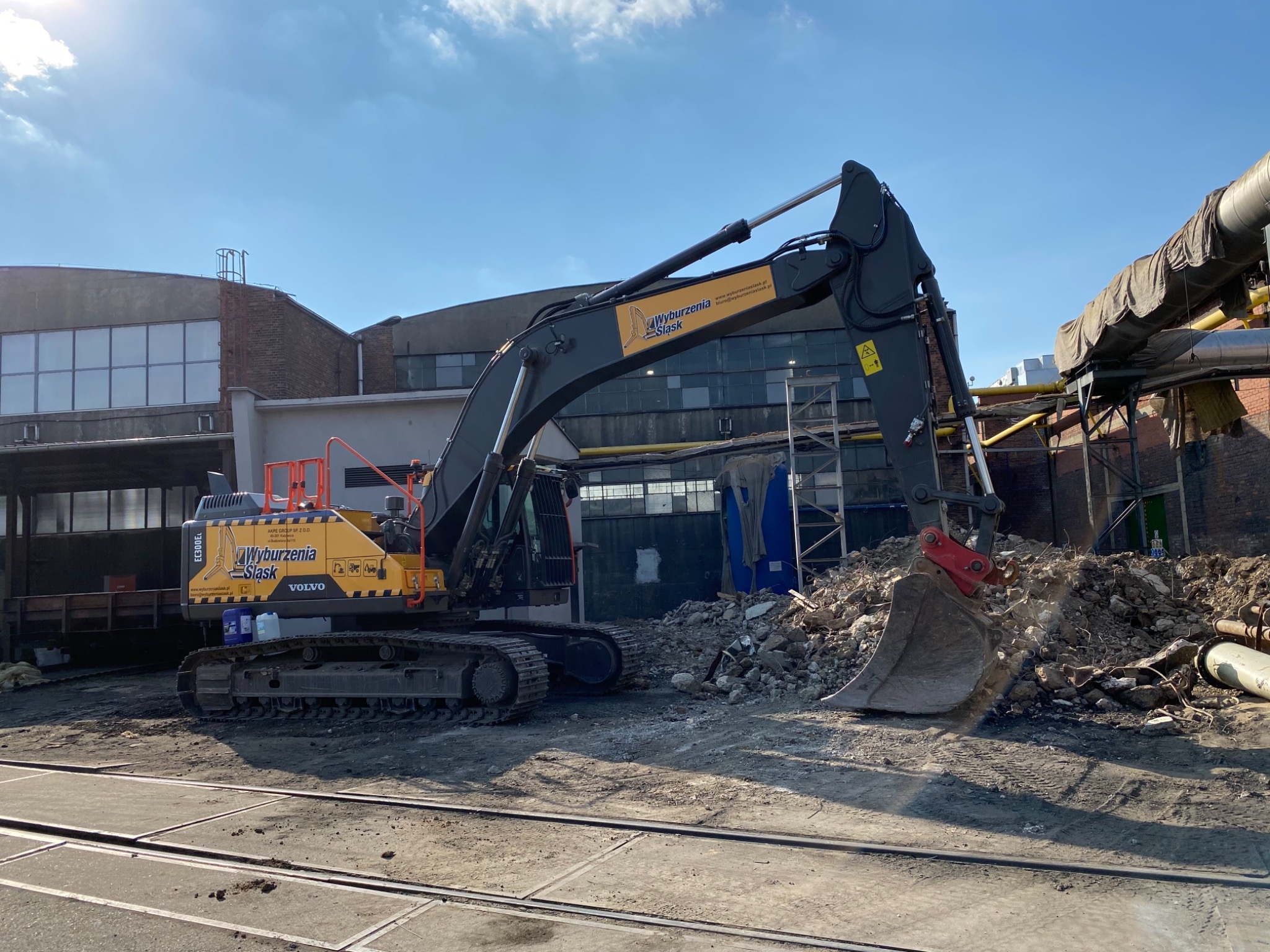 Żółto-czarna koparka Volvo z logo firmy 'Wyburzenia Śląsk' na tle gruzowiska i przemysłowego budynku z cegły