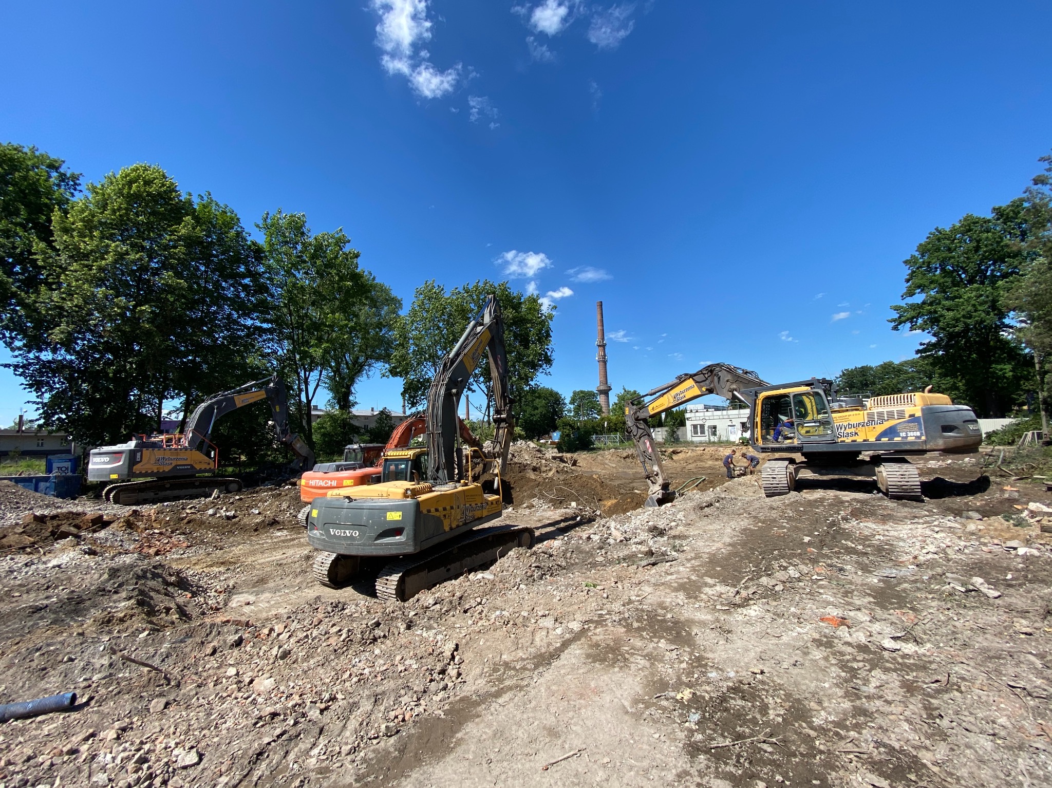 Trzy koparki Volvo i Hitachi na placu budowy w trakcie prac ziemnych, widoczny gruz i wykop, w tle drzewa i komin przemysłowy pod błękitnym niebem.