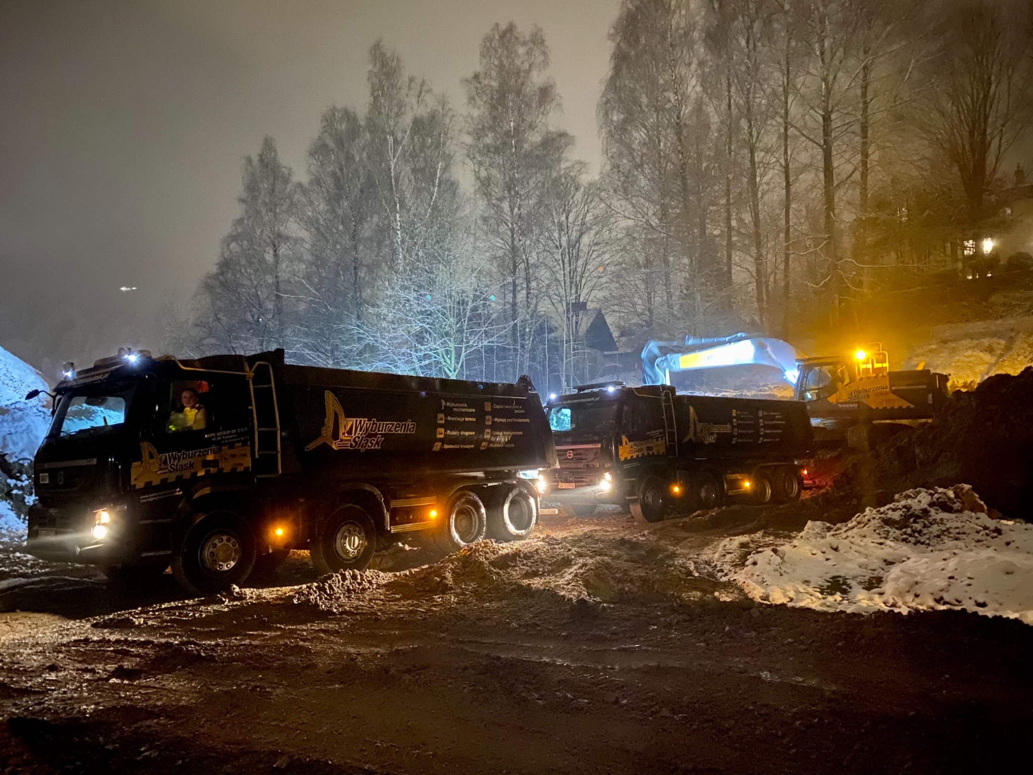 Dwa czarne samochody ciężarowe z logo firmy 'Wyburzenia Śląsk' stoją na błotnistym terenie w nocy, w tle widać koparkę i ośnieżone drzewa.
