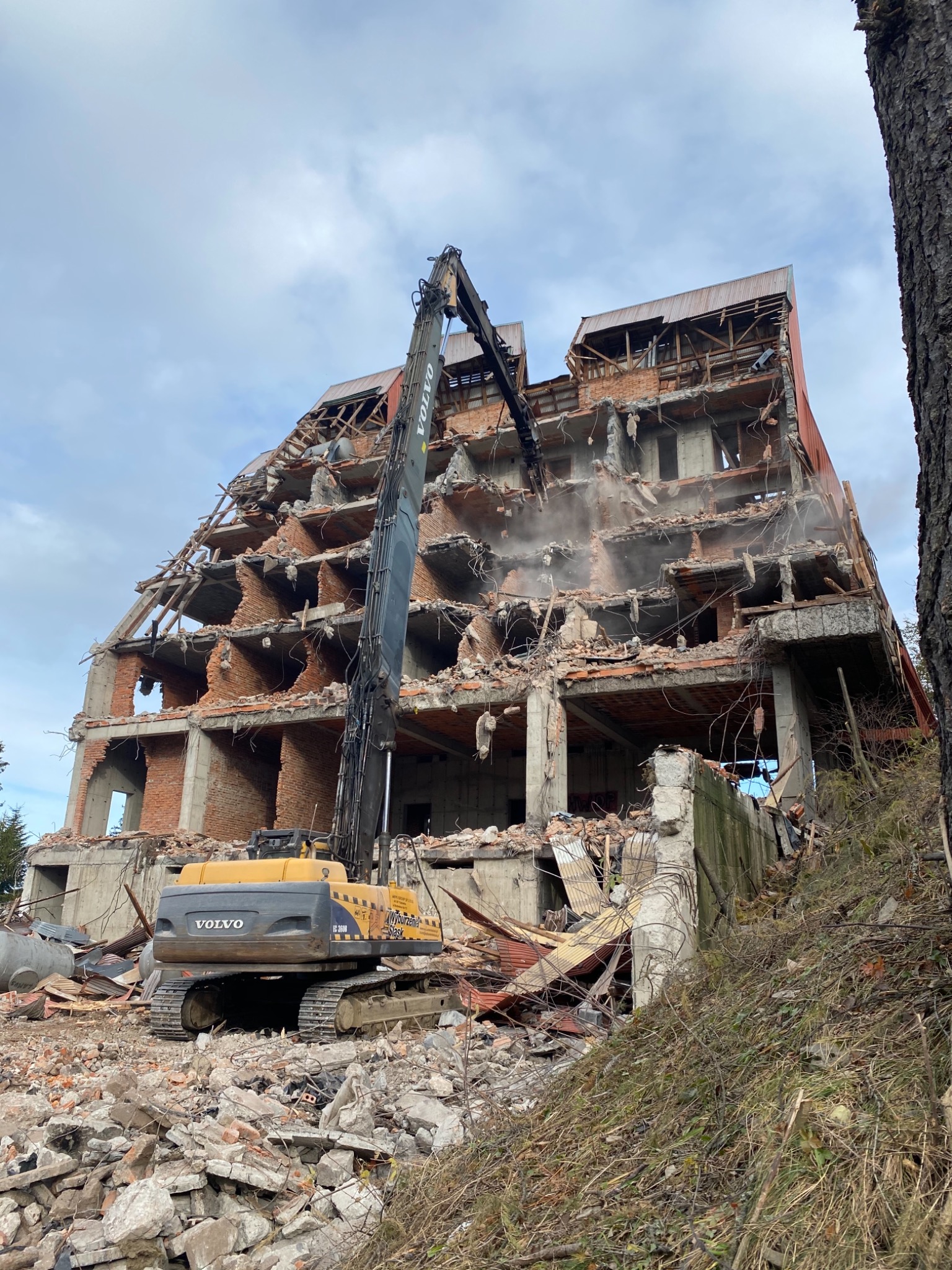 Żółto-szara koparka Volvo EC360B z długim ramieniem kruszy rozpadający się, wielopiętrowy budynek z czerwonej cegły i betonu, z widocznymi elementami drewnianej konstrukcji dachu, na tle...