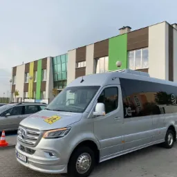 Srebrny bus Mercedes-Benz z logo DL-TRANS i naklejką ostrzegawczą dla dzieci, zaparkowany na tle kolorowego budynku szkoły.