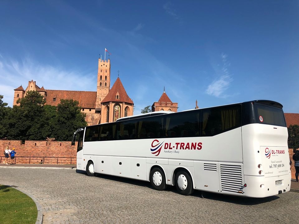 Biały autokar firmy DL-Trans z logo i numerem telefonu na tle ceglanego zamku w Malborku, widok z boku na kostce brukowej.