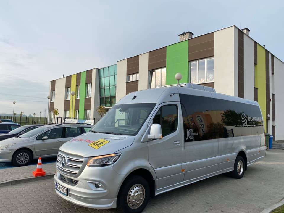 Srebrny bus Mercedes-Benz z logo DL-TRANS i naklejką ostrzegawczą dla dzieci, zaparkowany na tle kolorowego budynku szkoły.