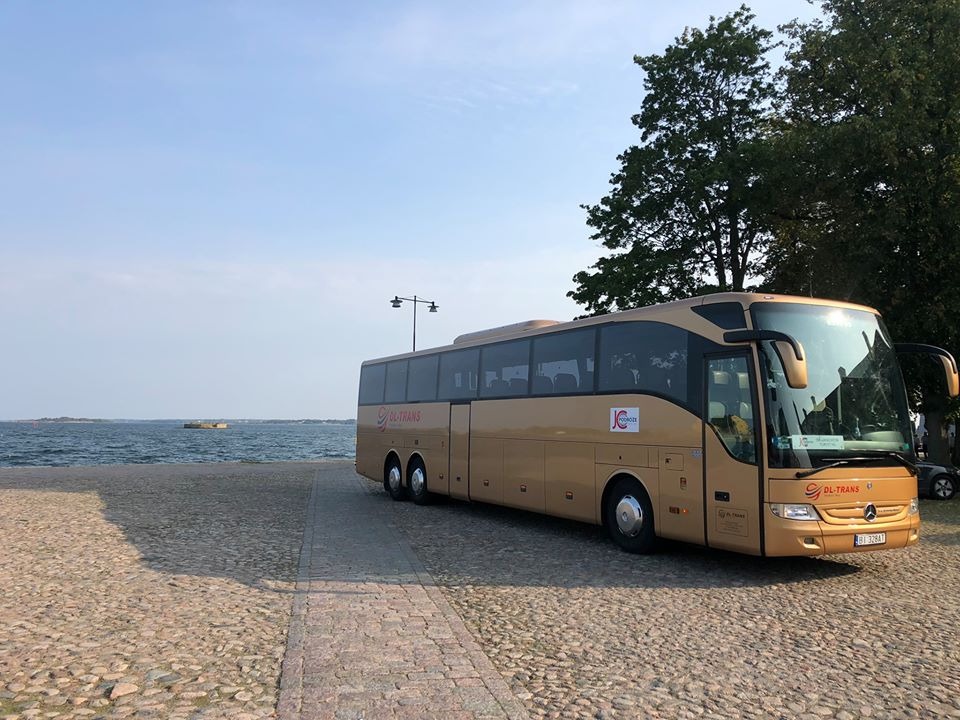 Złoty autokar turystyczny DL-Trans na tle morza i brukowanego nabrzeża, widoczny fragment drzewa i oświetlenie uliczne.
