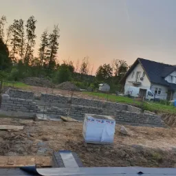 Budowa domu jednorodzinnego: fundamenty z izolacją, ściany fundamentowe z bloczków betonowych, paleta materiałów budowlanych w folii, w tle dom w trakcie budowy i zachodzące słońce.