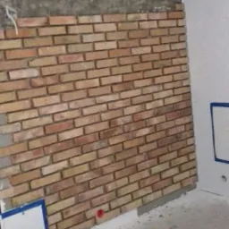 Montaż dekoracyjnej ściany z cegieł wewnątrz pomieszczenia, widoczne niedokończone górne partie oraz narzędzia i materiały budowlane na podłodze.