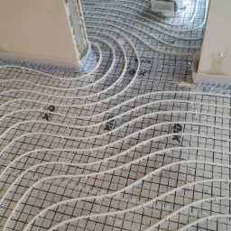 Białe rury ogrzewania podłogowego ułożone w pętle na siatce z tworzywa, widoczne zabezpieczenie piankowe przy ścianach, przygotowanie do wylewki.