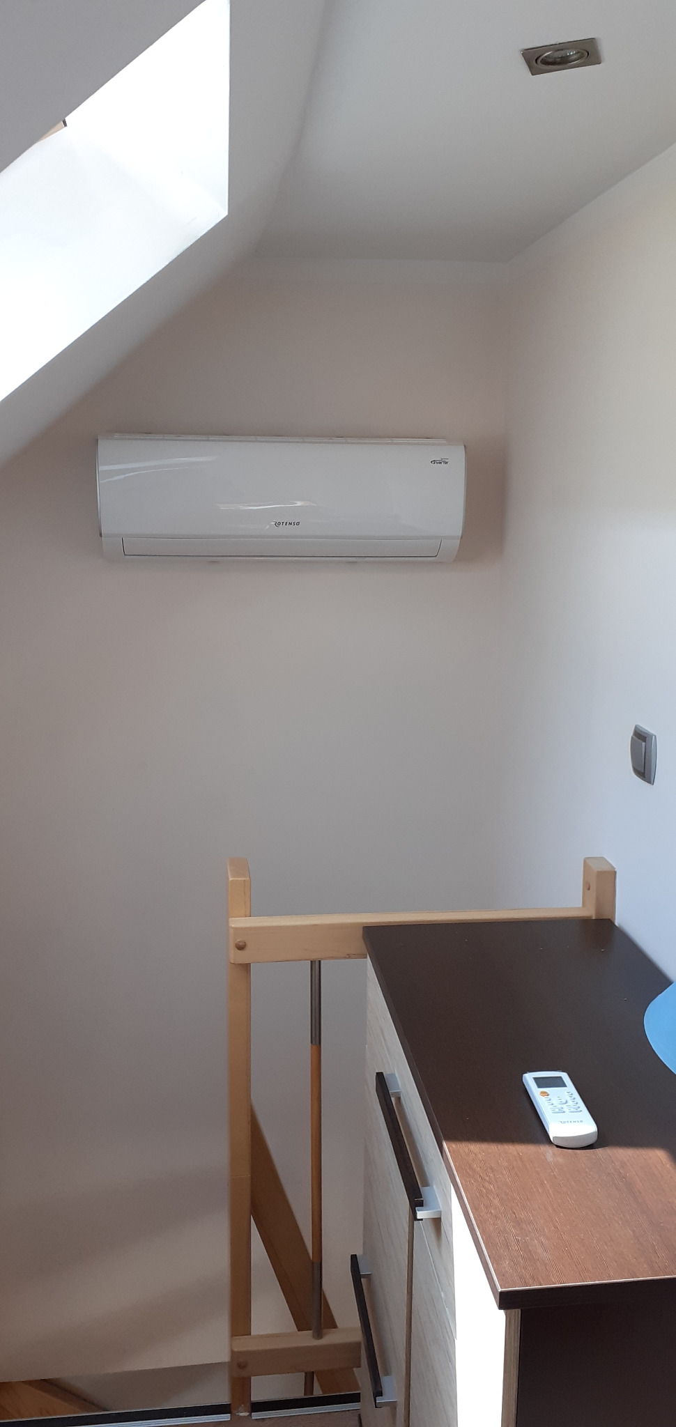 Biała jednostka wewnętrzna klimatyzatora Rotenso zamontowana na ścianie pod skosem dachu, widoczny fragment schodów i szafki z pilotem.