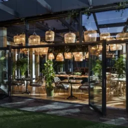 Nowoczesna restauracja z przeszkloną fasadą i składanymi drzwiami, oświetlona wiszącymi lampami z rattanu, z widocznymi stolikami i krzesłami wewnątrz.