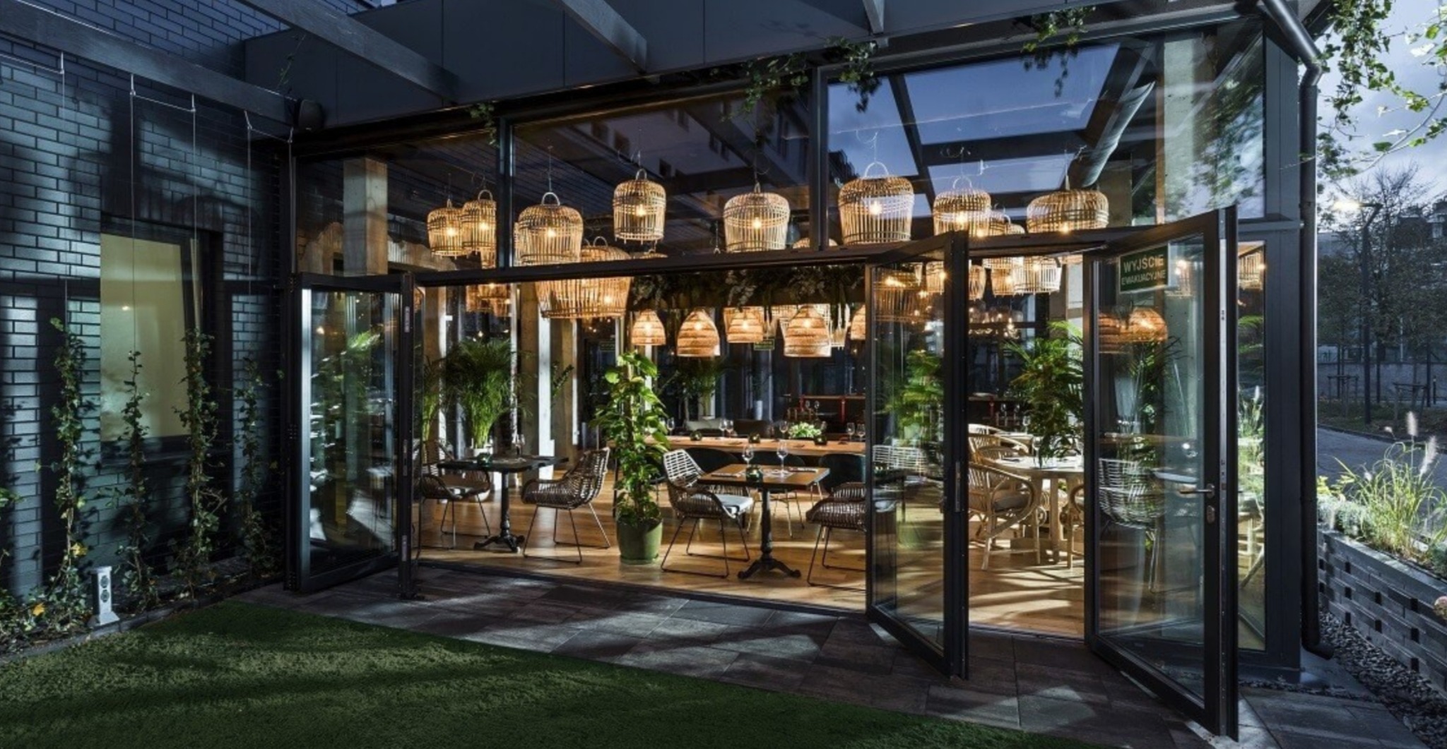Nowoczesna restauracja z przeszkloną fasadą i składanymi drzwiami, oświetlona wiszącymi lampami z rattanu, z widocznymi stolikami i krzesłami wewnątrz.