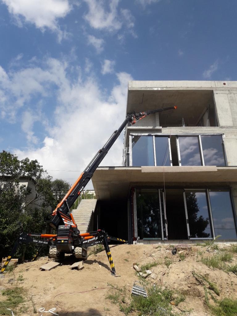 Montaż dużych okien aluminiowych w surowym stanie budynku za pomocą specjalistycznego dźwigu z przyssawkami, widoczne błękitne niebo z chmurami.