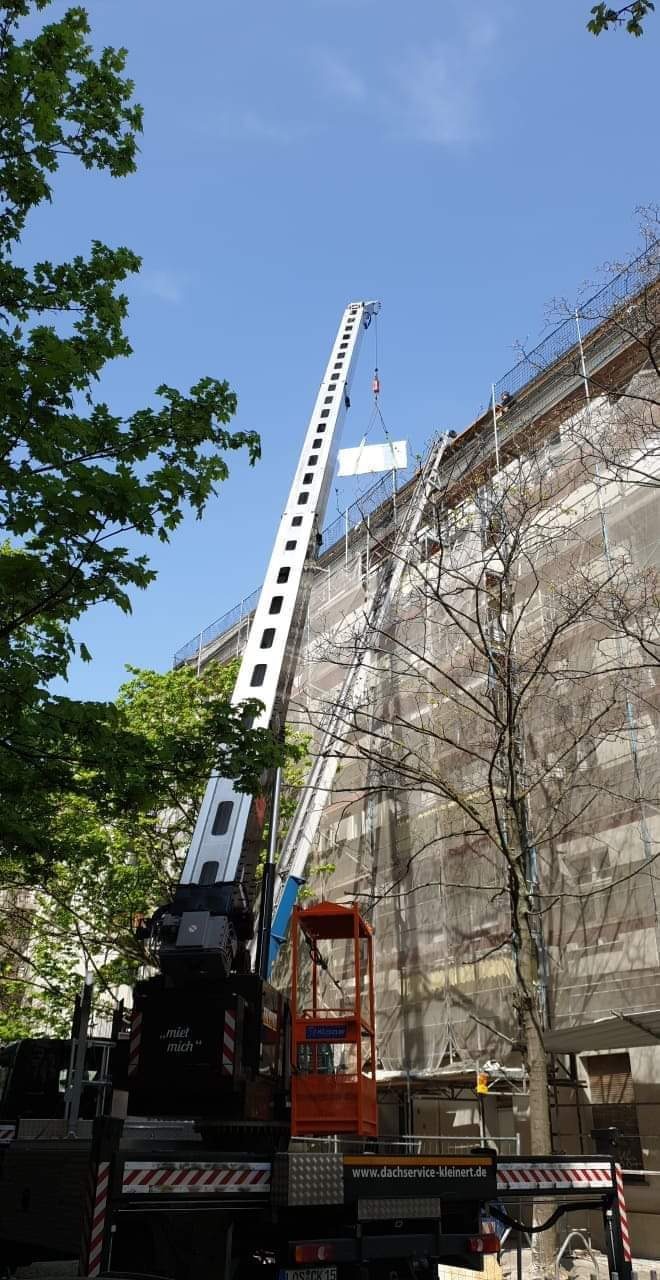 Podnośnik z platformą transportuje płytę na rusztowanie budynku podczas prac remontowych, widok z dołu na tle błękitnego nieba.
