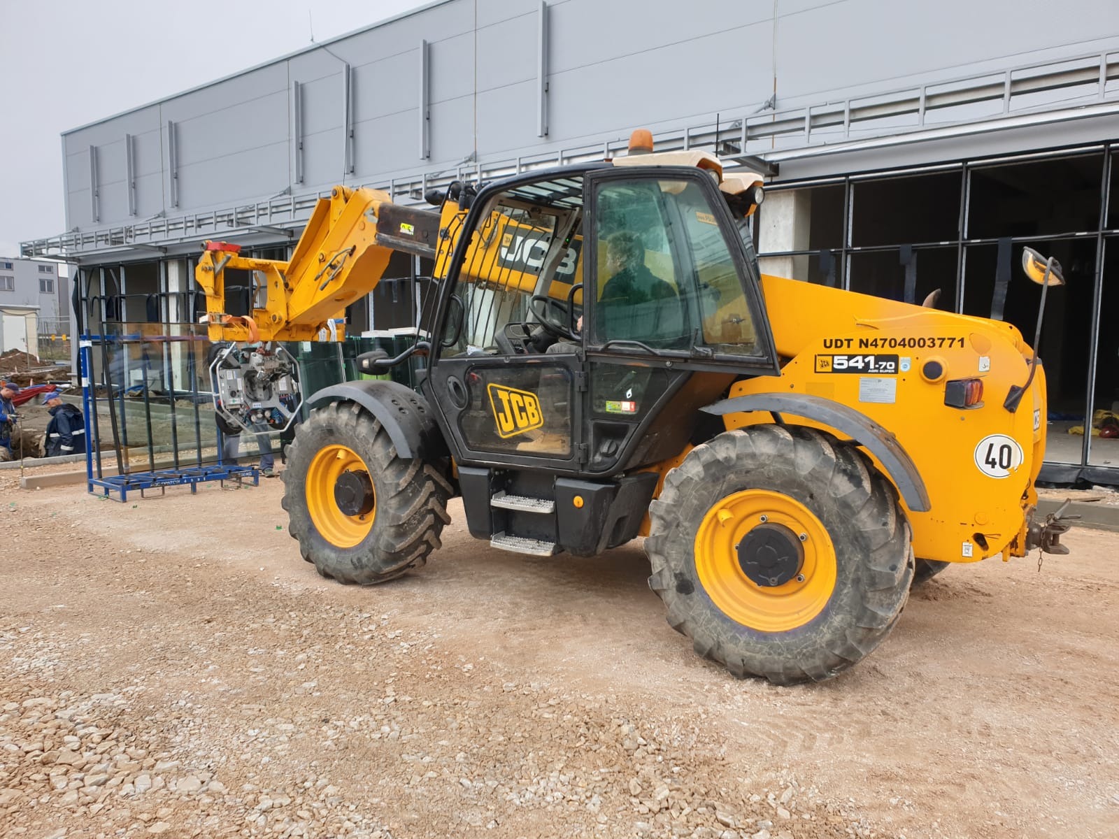 Żółta ładowarka teleskopowa JCB podnosi szybę na placu budowy, widoczni pracownicy w tle.