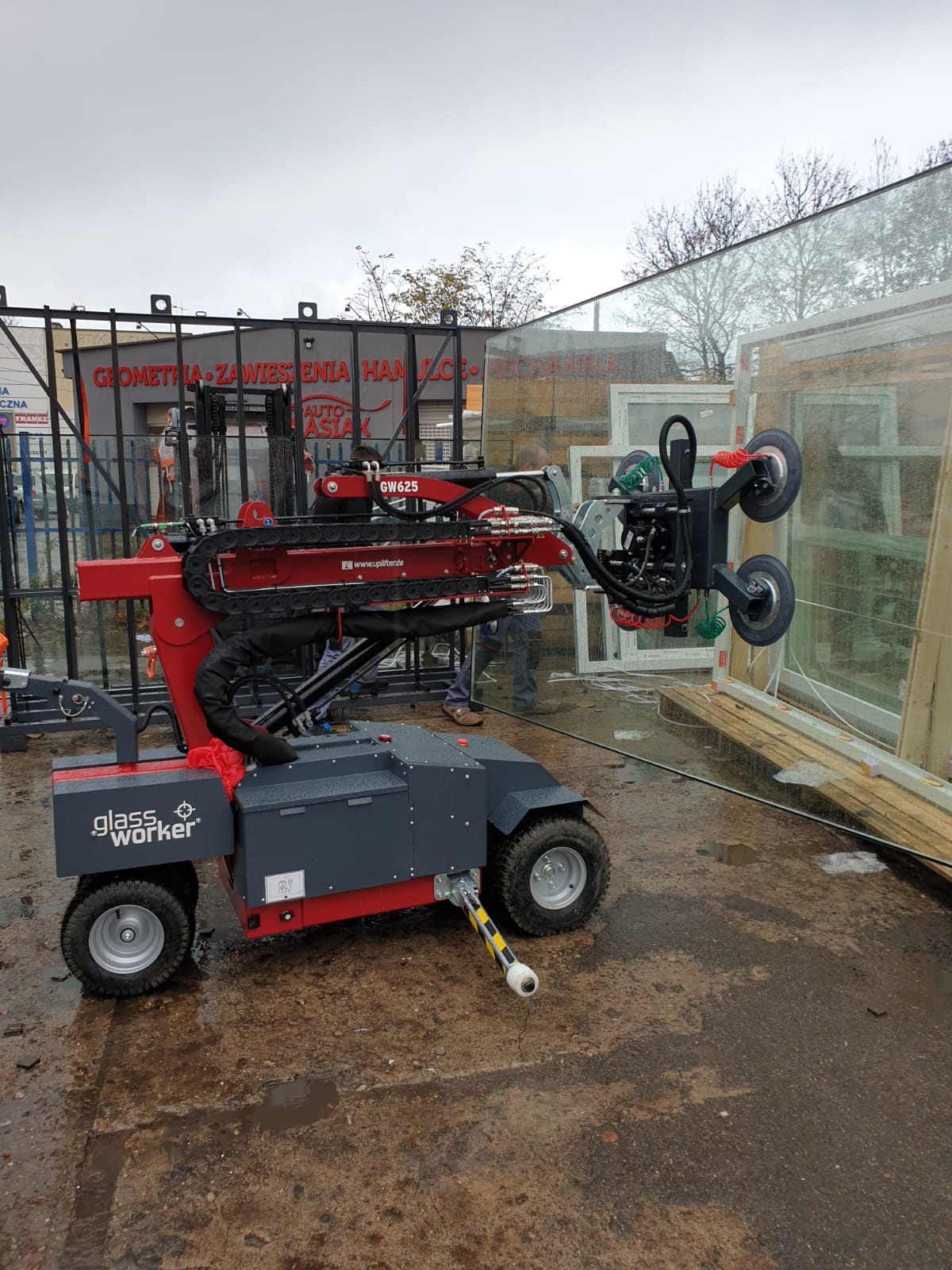 Maszyna do przenoszenia szyb Glassworker GW625 z przyssawkami, unosząca dużą szybę na zewnątrz budynku, w tle składowane ramy okienne.