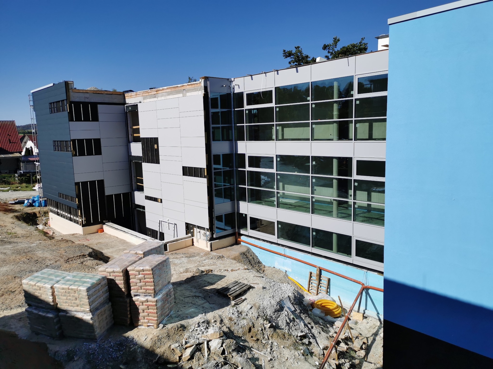 Wznoszony budynek z aluminiowymi oknami podczas prac wykończeniowych, widoczne worki z materiałami budowlanymi i teren budowy.