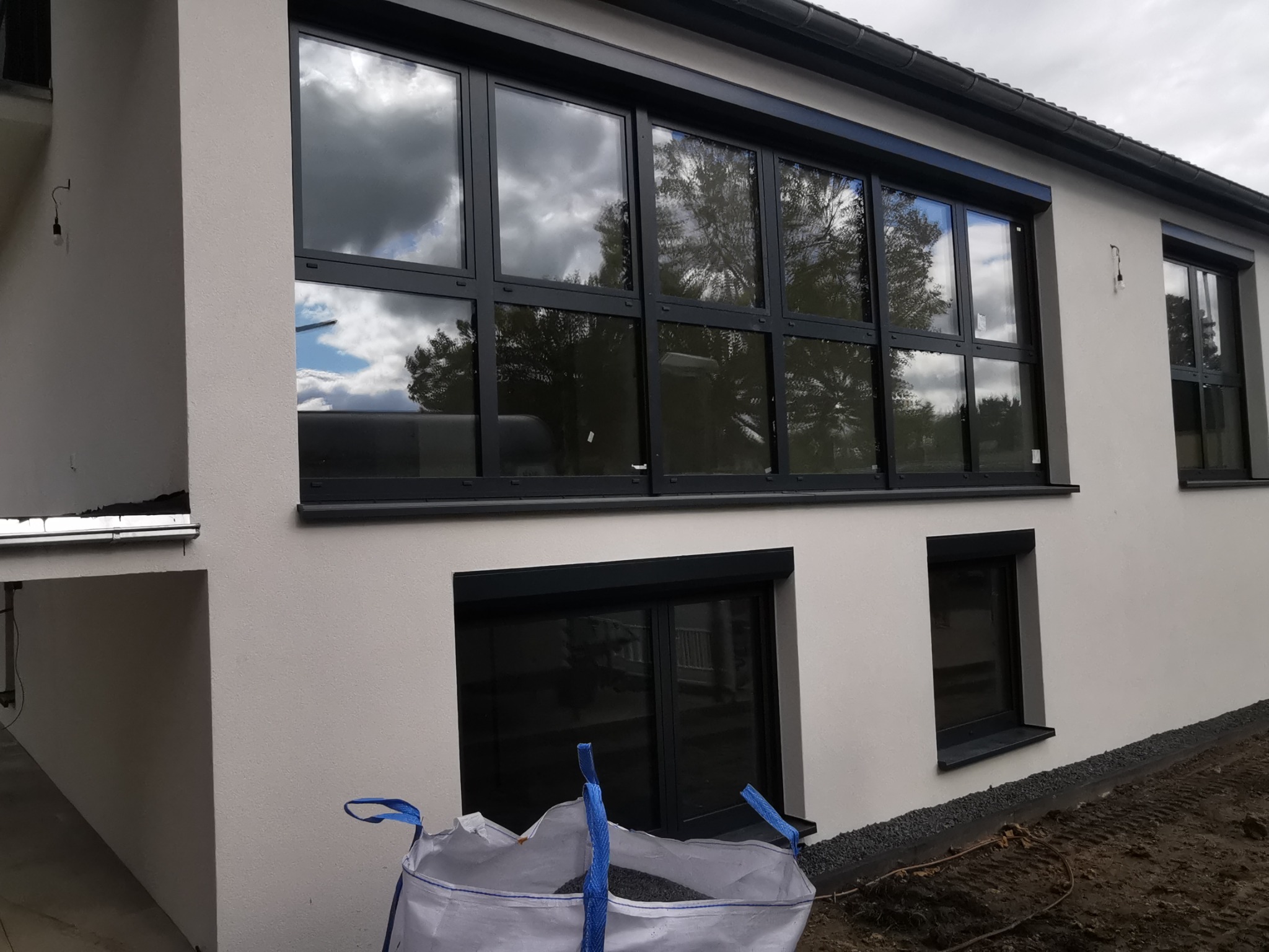 Nowoczesna fasada budynku z dużymi aluminiowymi oknami z czarnymi ramami, odbijającymi chmury i drzewa, z workiem wypełnionym materiałem budowlanym w dolnej części kadru.