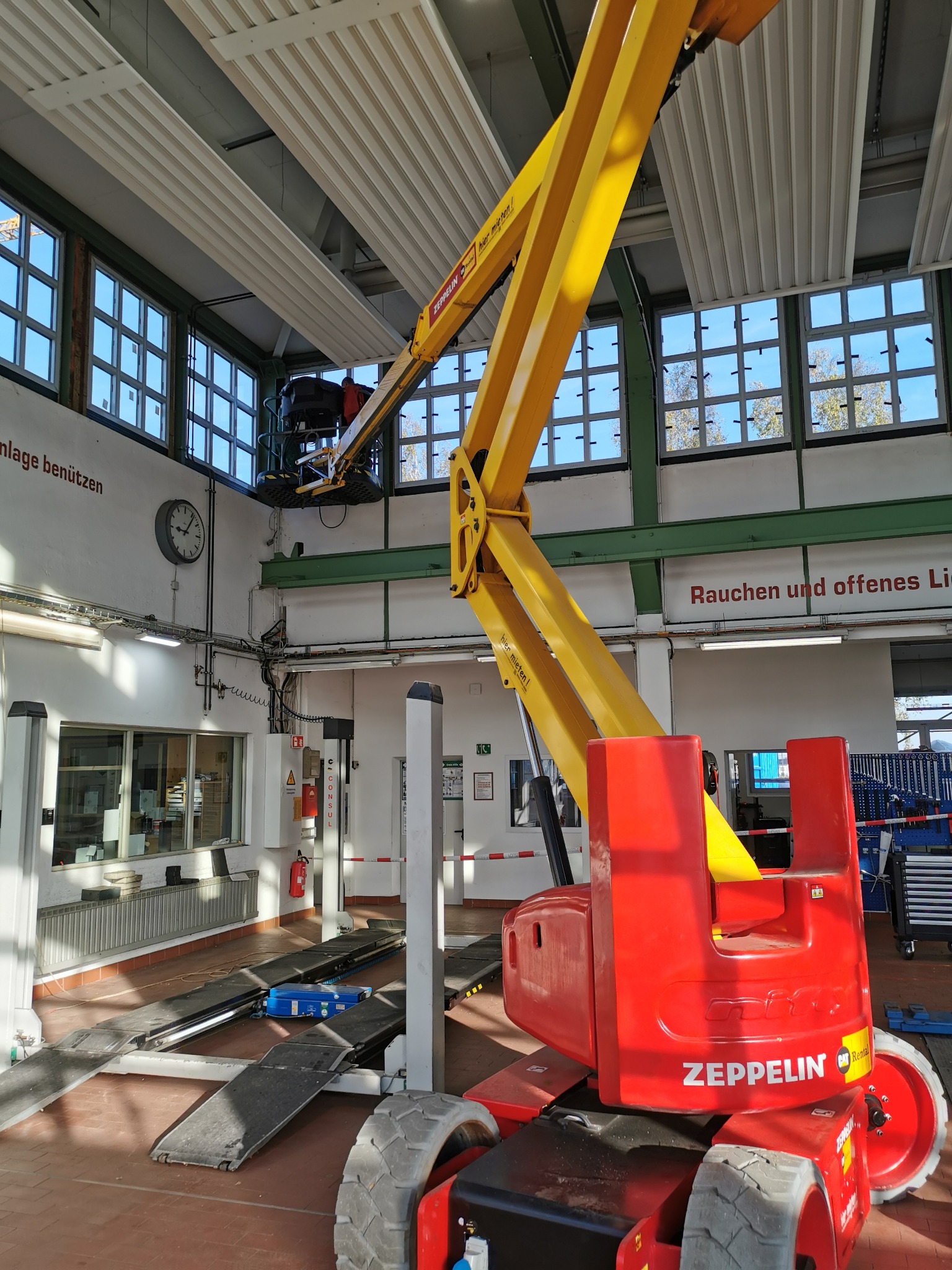 Czerwono-żółty podnośnik Zeppelin w hali z dużymi oknami, pracownik na podnośniku przy suficie z panelami.
