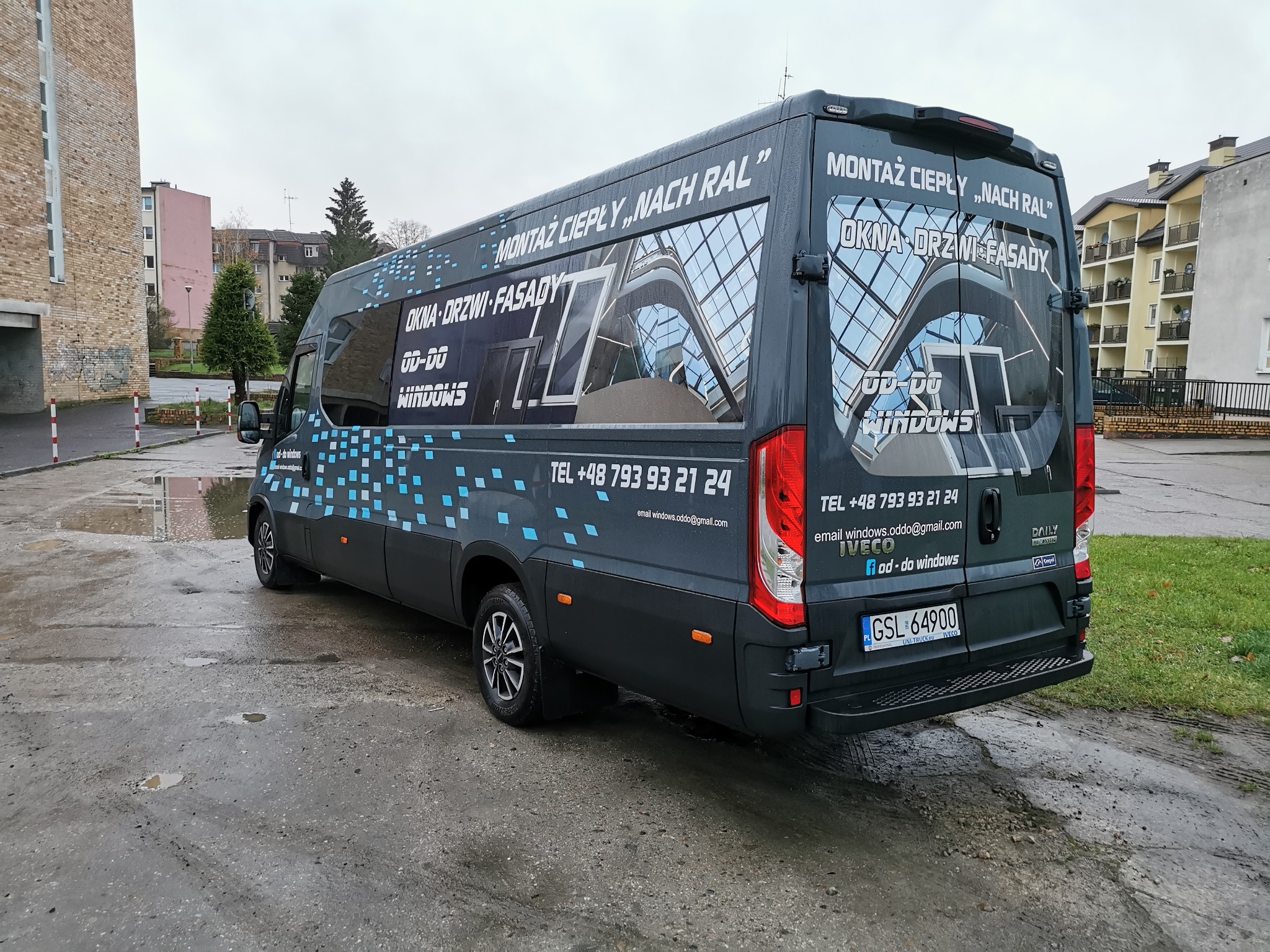 Czarny samochód dostawczy z reklamą firmy montującej okna, drzwi i fasady, zaparkowany na mokrym parkingu przed budynkami.