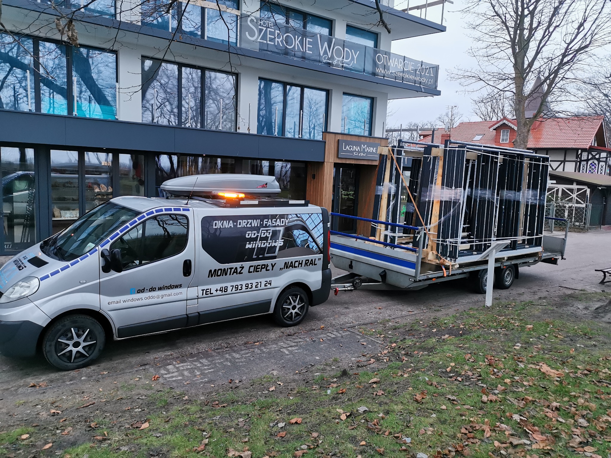 Srebrny samochód dostawczy z przyczepą wypełnioną pakietami nowych okien na tle budynku Hotelu Szerokie Wody, sugerujący dostawę i montaż stolarki okiennej.