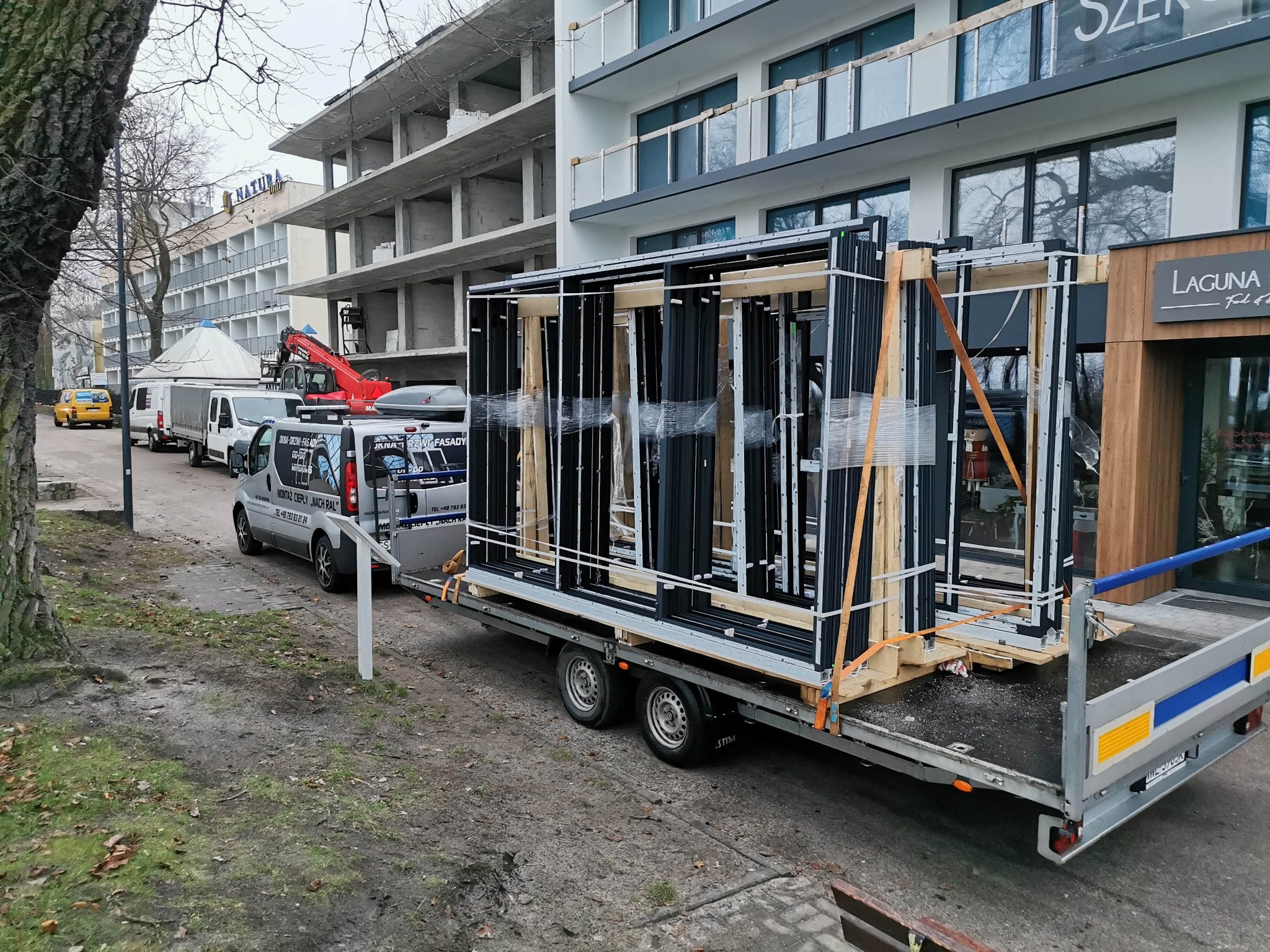 Transport okien aluminiowych na przyczepie, budynek w tle, widoczny samochód dostawczy z reklamą firmy.