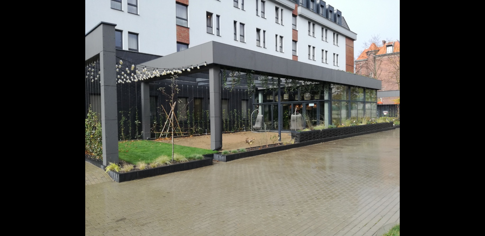 Nowoczesna pergola z oświetleniem łańcuchowym i zielenią, przylegająca do budynku, z widoczną kostką brukową.