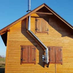 Zewnętrzny montaż komina ze stali nierdzewnej na drewnianym domku letniskowym z brązowymi okiennicami, widok z boku, słoneczny dzień.